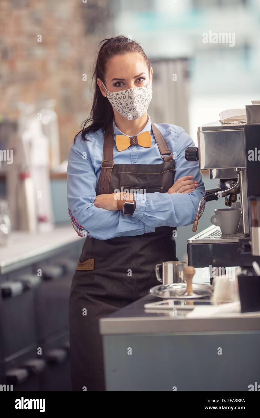 Gesichtsmaske trägt Kellnerin steht in einer Bar neben einer Kaffeemaschine mit Armen auf ihrer Brust ruhen. Stockfoto