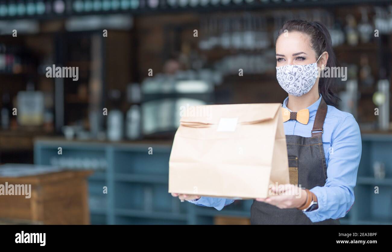 Restaurantmitarbeiter übergibt während der Pandemie von Covid-19 Lebensmittel, die in einer Papiertüte mit Gesichtsmaske verpackt sind. Stockfoto