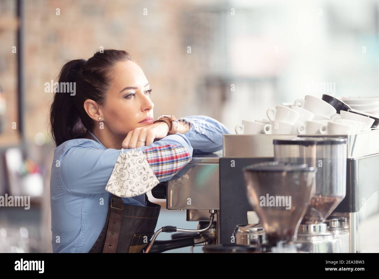 Gelangweilte Kellnerin lehnt sich an die Kaffeemaschine ohne Arbeit zu tun, hält Gesichtsmaske in der Hand. Stockfoto