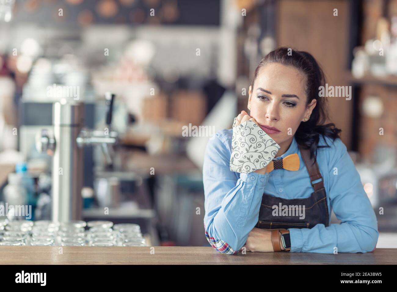 Traurige Geschäftsbesitzerin hält Gesichtsmaske in der Hand und lehnt sich an die Bar in ihrem leeren Café. Stockfoto