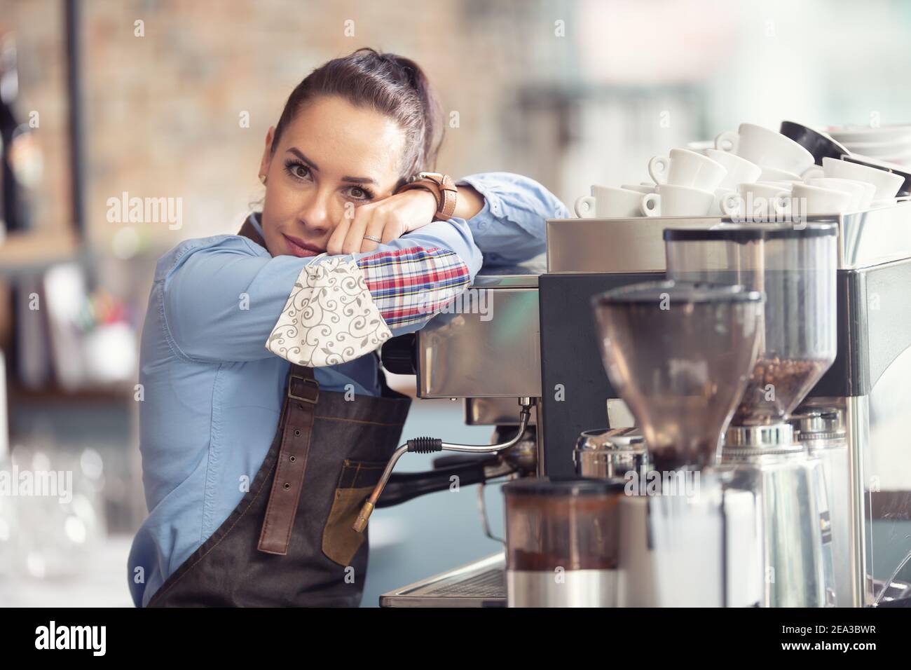 Gelangweilte Kellnerin lehnt sich an die Kaffeemaschine ohne Arbeit zu tun, hält Gesichtsmaske in der Hand. Stockfoto