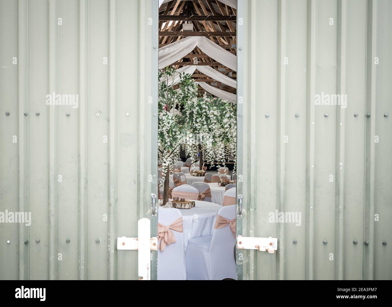 Schleichen einen Blick auf Hochzeitsempfangsfeier hinter gelegt Rustikale Türen in traditionellen alten umgebauten Bauernhof Scheune mit Blüte Bäume weiße Tische Stockfoto