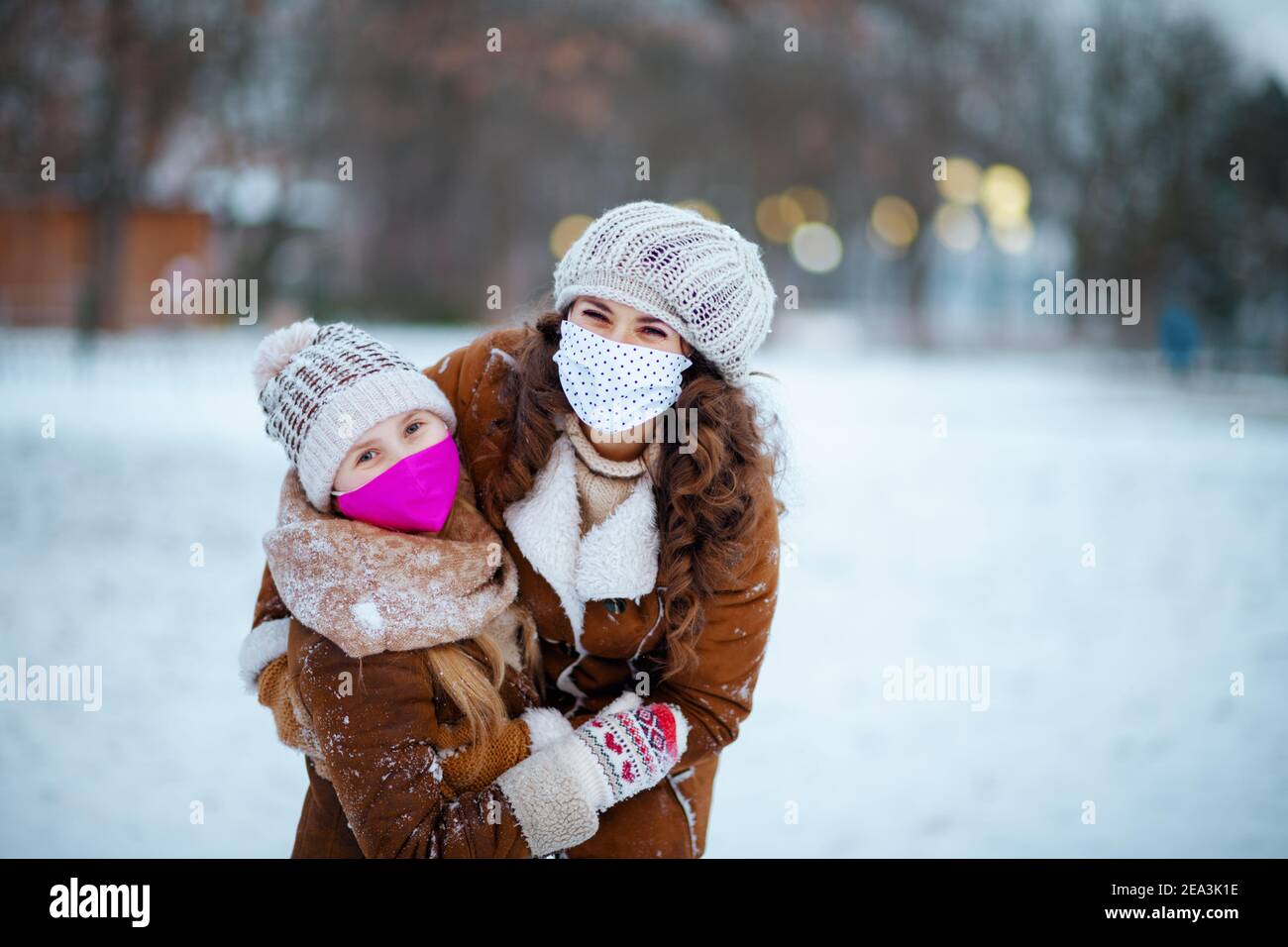 Porträt von lächelnd stilvolle Mutter und Kind in einem gestrickten Hüte und Schaffell Mäntel mit Fäustlingen und medizinische Maske in Eine Strickmütze und Schaffell Mantel o Stockfoto