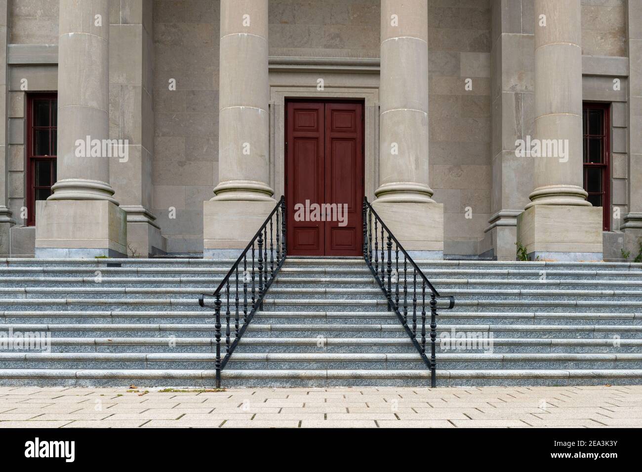 Sechs große runde Betonsäulen an der Spitze der Marmortreppe mit schwarzen Eisenschienen zu einem legalen Gebäude. Das Regierungsgebäude hat eine hohe rote Tür. Stockfoto