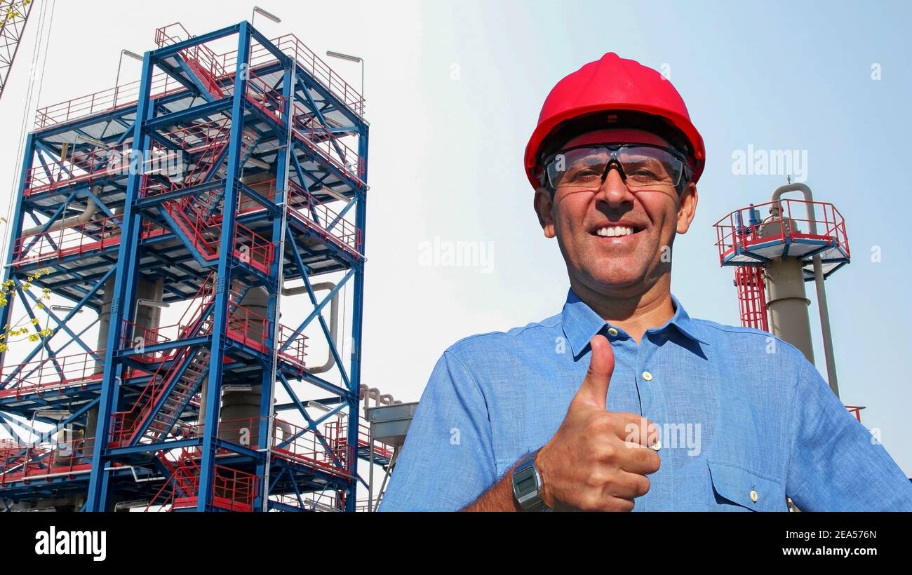 Happy Oil Worker in Red HardHat gibt Daumen nach oben vor Öl und Gas Raffinerie. Porträt eines lächelnden Mannes in schützender Arbeitskleidung. Stockfoto