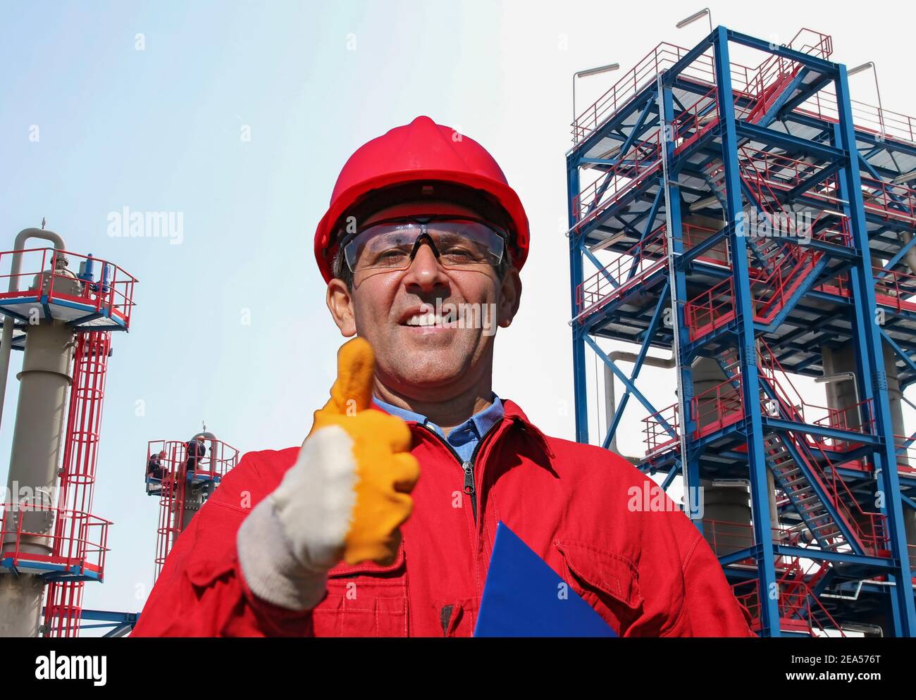 Happy Oil Worker in Red HardHat gibt Daumen nach oben vor Öl und Gas Raffinerie. Porträt eines lächelnden Mannes in schützender Arbeitskleidung. Stockfoto