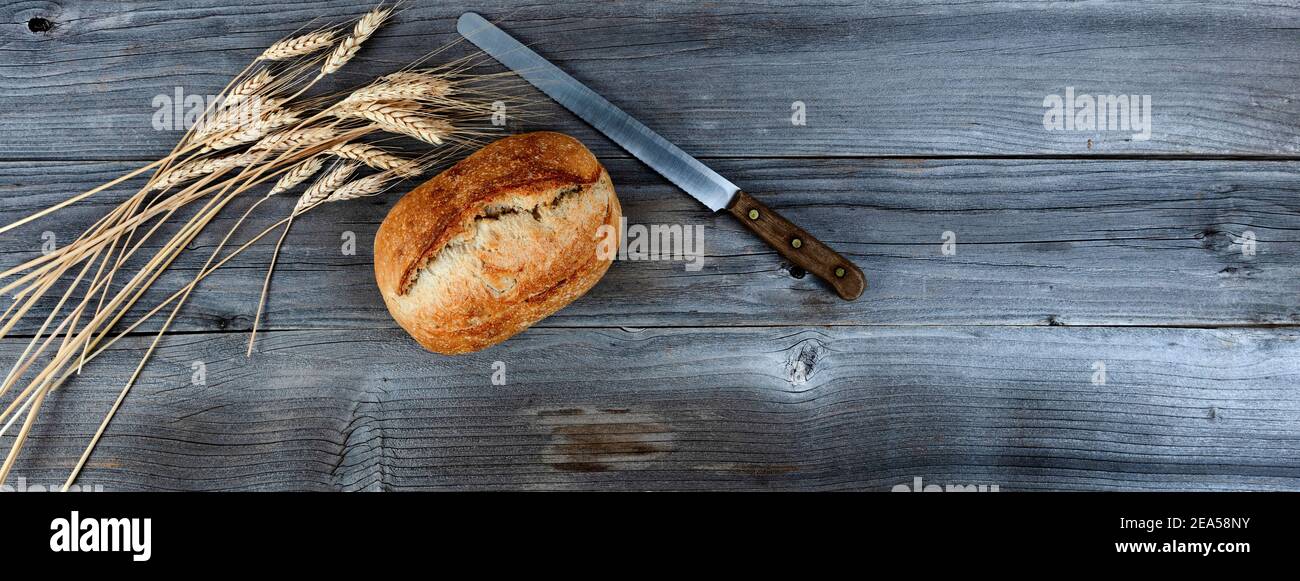 Blick von oben auf hausgemachtes frisch gebackenes Brot mit Schneidemesser und getrocknete Weizenstiele auf verwitterten Holzbohlen Stockfoto