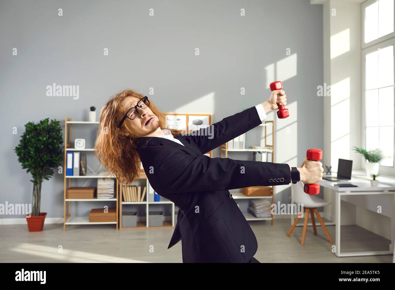 Lustige nerdy Mann im Büro Anzug versuchen, mit auszuüben Kurzhanteln während des Arbeitstages Stockfoto