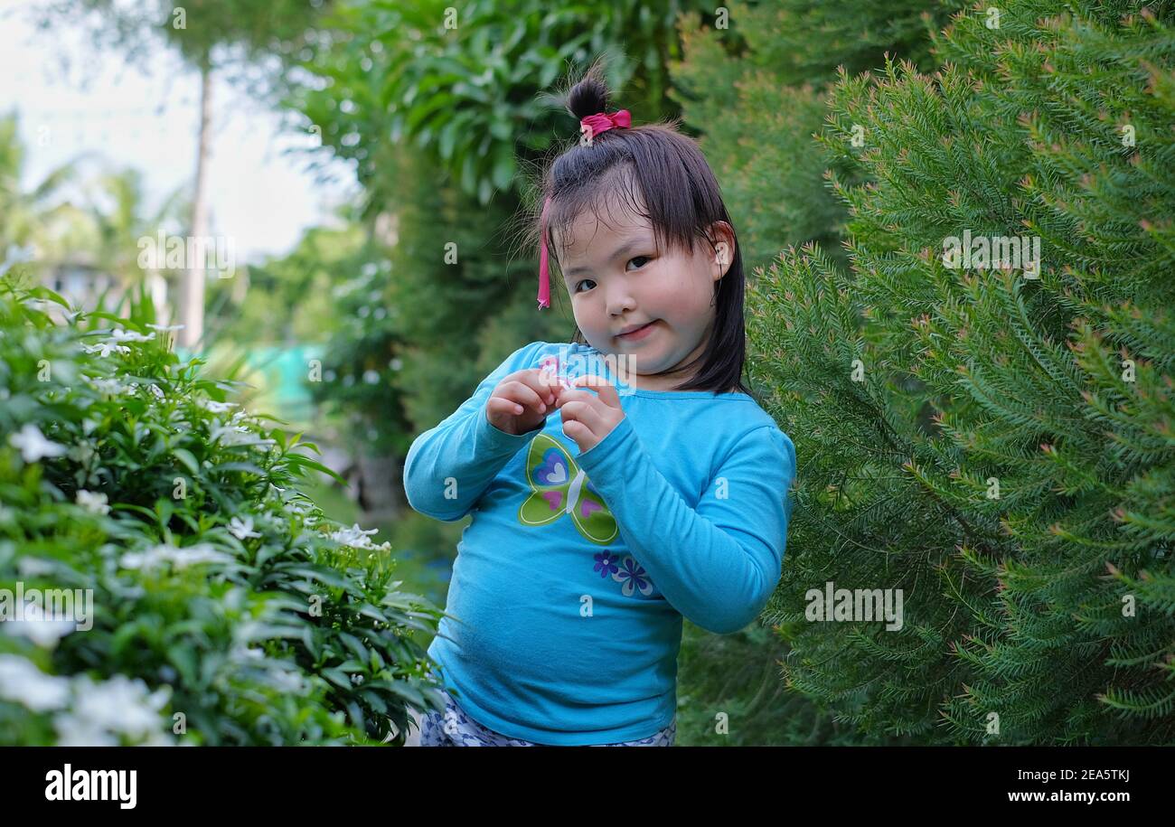 Ein süßes junges asiatisches Mädchen posiert für die Kamera in einem Garten mit Bäumen im Hintergrund, lächelnd, aber schüchtern. Stockfoto