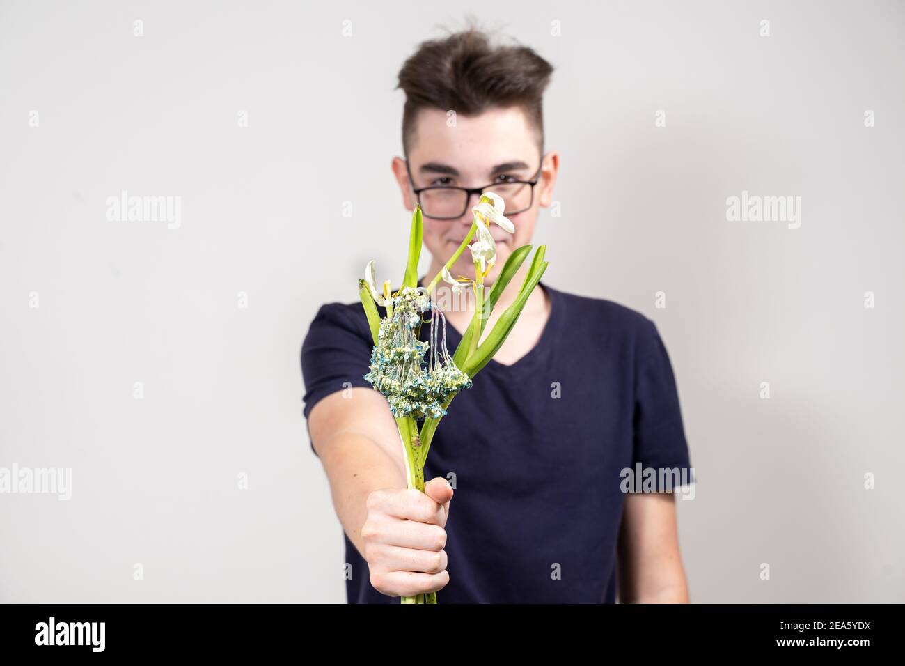 Ein junger Mann mit Brille hält einen Strauß von verwelkt Blumen und lächelt. Das Konzept der Gier und Geiz. Konzentrieren Sie sich auf Blumen, Kerl in Unschärfe Stockfoto