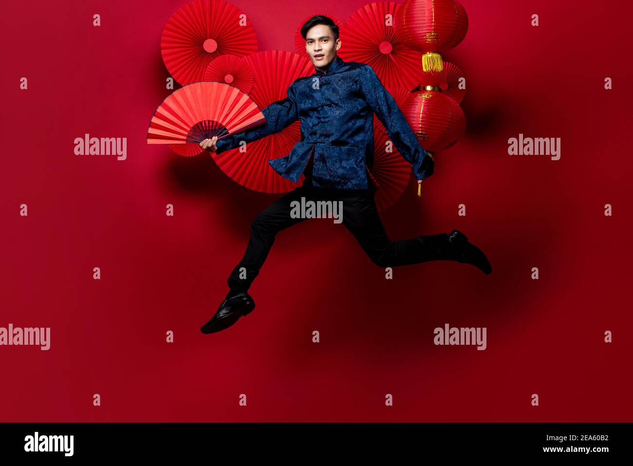 Asiatischer Mann in traditioneller Tracht hält Fan und springen in orientalischer Stil dekoriert roten Hintergrund für chinesische Neujahrs-Konzepte Stockfoto