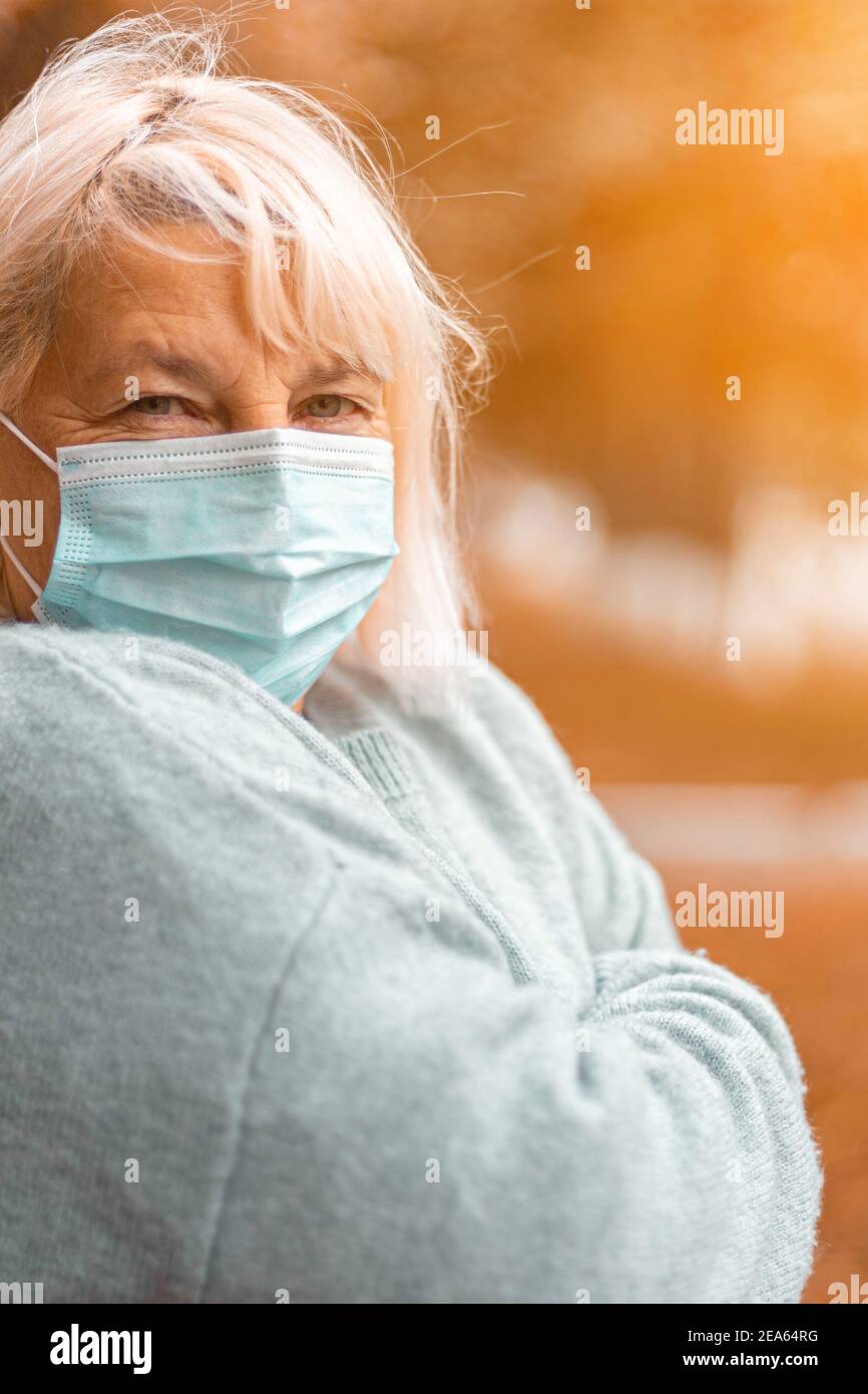 Blonde kaukasische Frau 50 Jahre alt in einem gemütlichen warm Pullover und eine medizinische Schutzmaske, die auf die Kamera schaut In einem Herbstpark Stockfoto