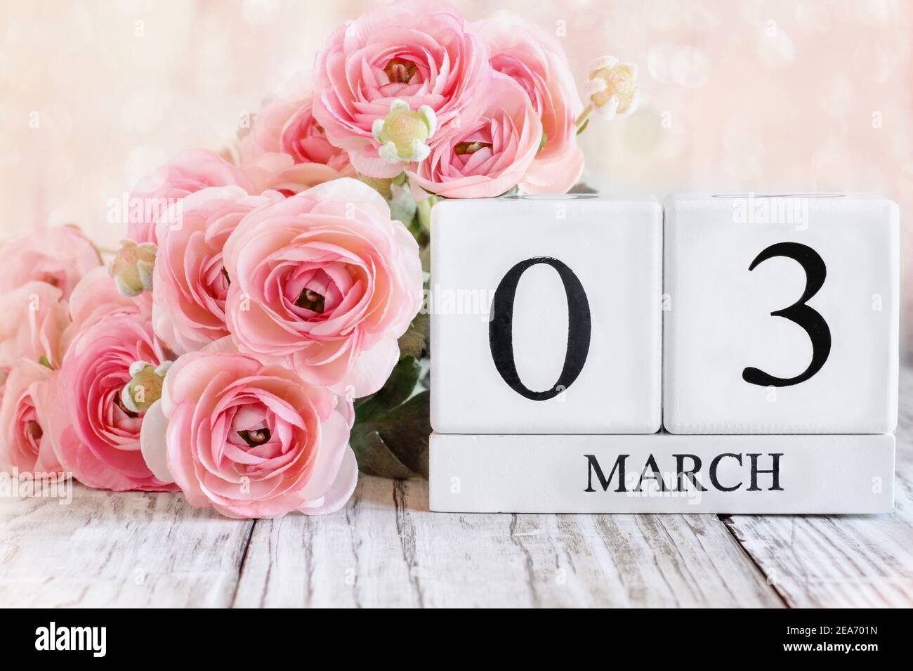 Weiße Holz-Kalenderblöcke mit dem Datum März 3rd und rosa Ranunculus Blumen über einem Holztisch. Selektiver Fokus mit unscharfem Hintergrund. Stockfoto