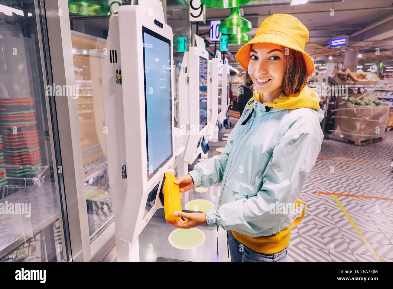 Glückliche junge Kundin Frau kauft und zahlt für eine Flasche Orangensaft an der Kasse eines SB-Automaten in einem modernen Supermarkt, um zu AV Stockfoto