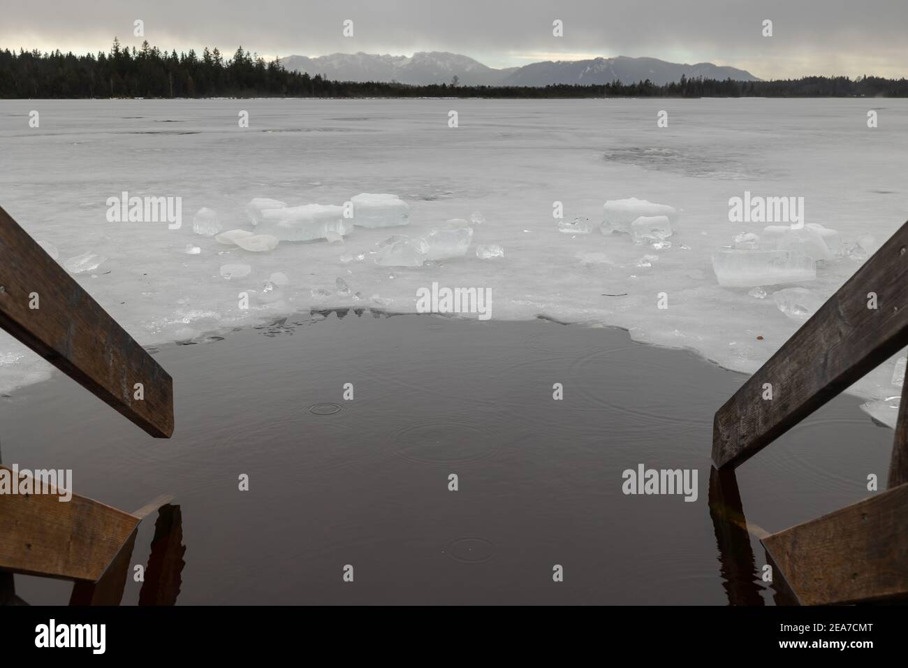 Loch im Eis zum Baden neben hölzernem Nasenstück auf gefroren see in Oberbayern mit Alpen im Hintergrund im Winter Mit Schnee Stockfoto