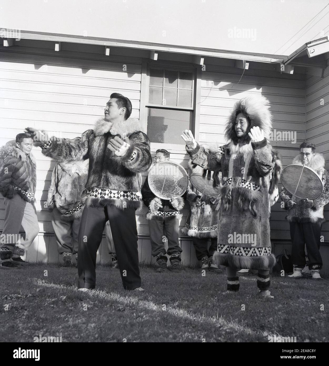 1960, historisch, an der Außengrenze von Anchorage, Alaska, USA, außerhalb eines einstöckigen Holzgebäudes, Ureinwohner in traditioneller Kleidung aus Tierfellen und Pelzen, die einen Tanz aufführen. Alaska wurde 49th der Staat der USA im Jahr 1959, aber es wurde von verschiedenen indigenen Völkern für Tausende von Jahren vor der Ankunft der Europäer besetzt. Da der Staat eine Seegrenze zu Russland hat, waren es sie, die sich als erste niederließen, was als russisches Amerika bekannt wurde. Die indigenen Völker machen über 15% der Bevölkerung aus und die alaskischen Ureinwohner spielen eine wichtige Rolle in der lokalen Regierung Stockfoto