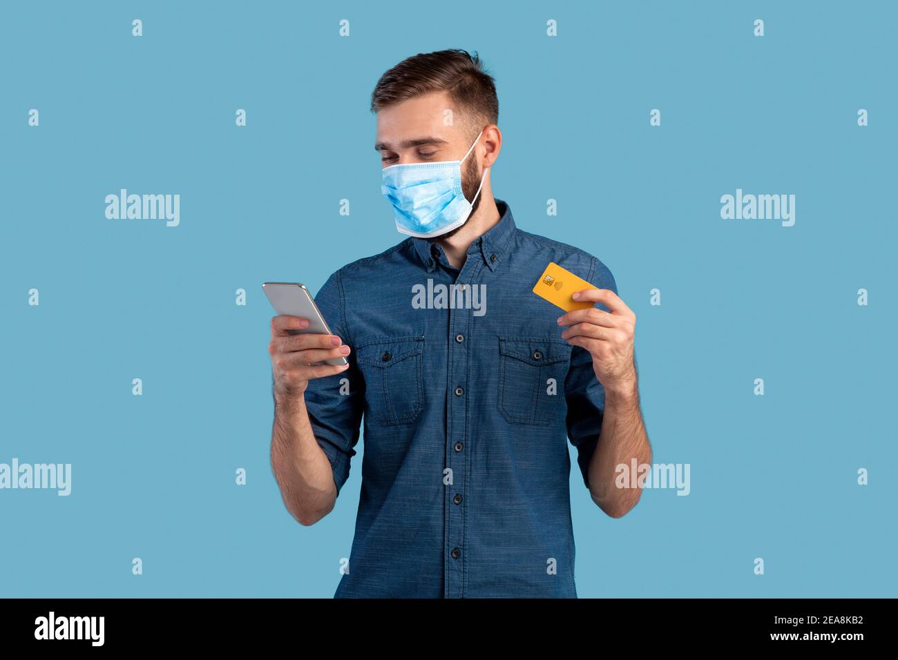 Kontaktloses Einkaufen während der Covid-Isolation. Porträt von Millennial Kerl in Gesichtsmaske hält Smartphone und Kreditkarte Stockfoto