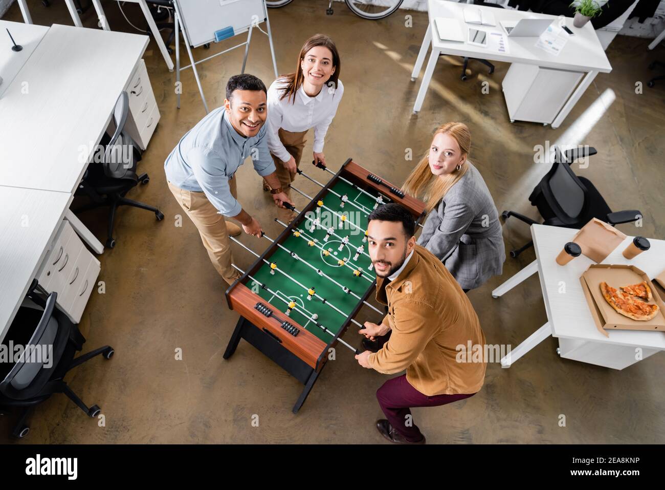 Blick von oben auf fröhliche, multiethnische Geschäftsleute, die Tischfußball spielen In der Nähe von Pizza im Büro Stockfoto