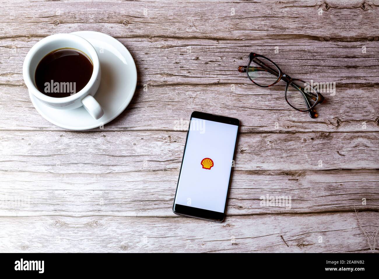 Ein Handy oder Handy auf einem Holz gelegt Tisch mit der Shell-App neben einem Kaffee geöffnet Und Brille Stockfoto