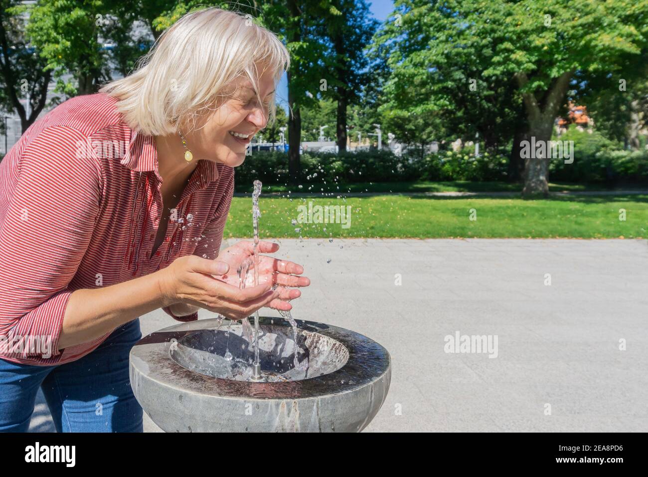 Reife Frau erfrischend aus Trinkwasser Brunnen am heißen Sommertag. Blonde Frau spritzt Wasser von Brunnen. Frisch durch die Hitze Stockfoto