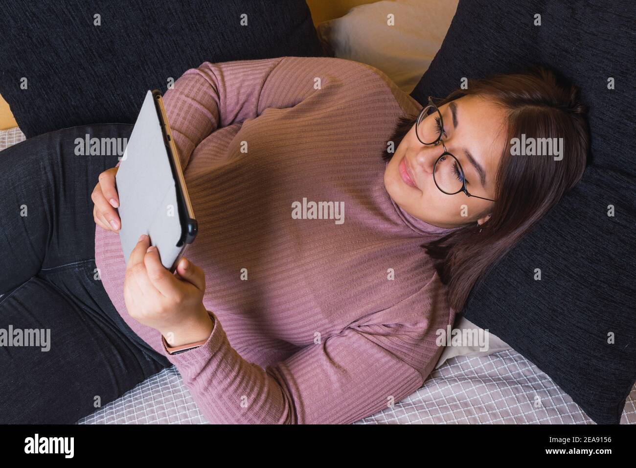 Junge lateinische Frau streckte sich auf ihrem Schlafbett aus Ihr Kopf auf einem blauen Kissen und schaute auf das Tablet Stockfoto