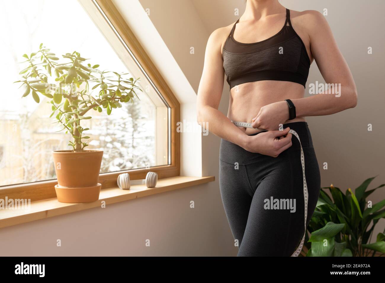 Junge schöne Frau Messung Taille mit Band durch ein Fenster im Dachboden an sonnigen Tag. Gesundes Wohnen und Training zu Hause. Stockfoto