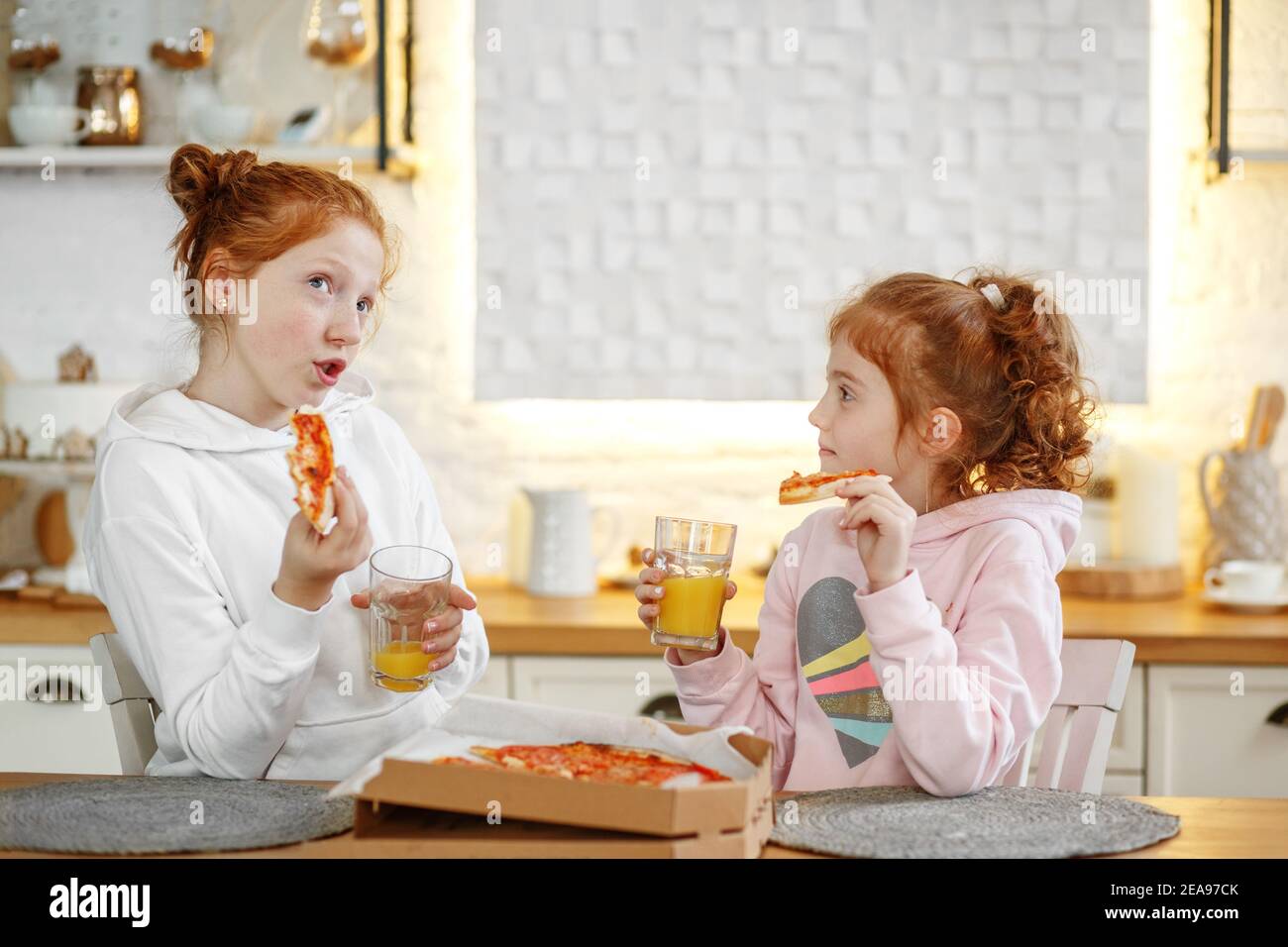 Junge Mädchen mit roten Haaren in der Küche essen Pizza, trinken Saft. Treffen mit Freundinnen, zwei Schwestern. Stockfoto