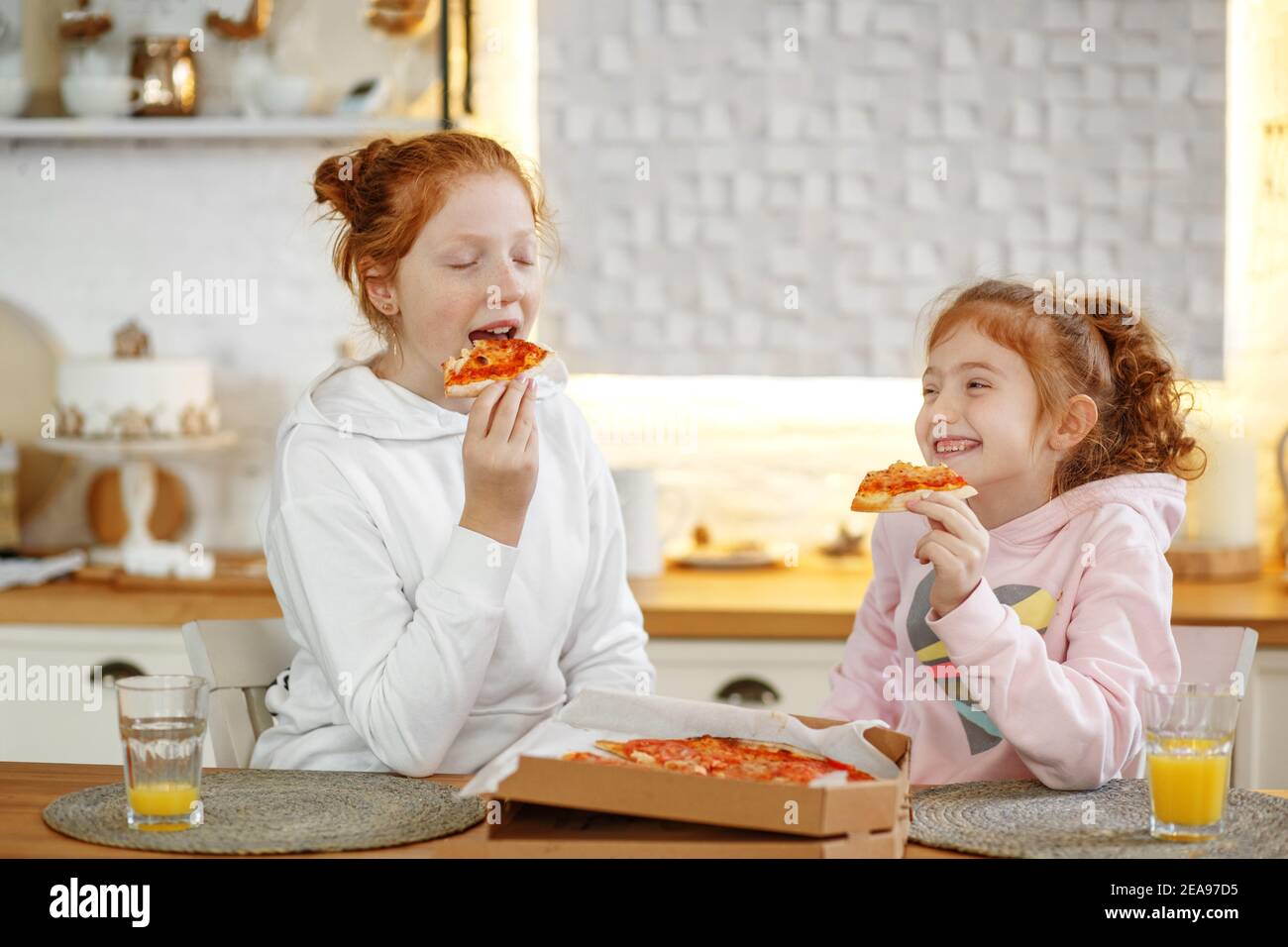 Junge Mädchen mit roten Haaren in der Küche essen Pizza, trinken Saft. Treffen mit Freundinnen, zwei Schwestern. Stockfoto