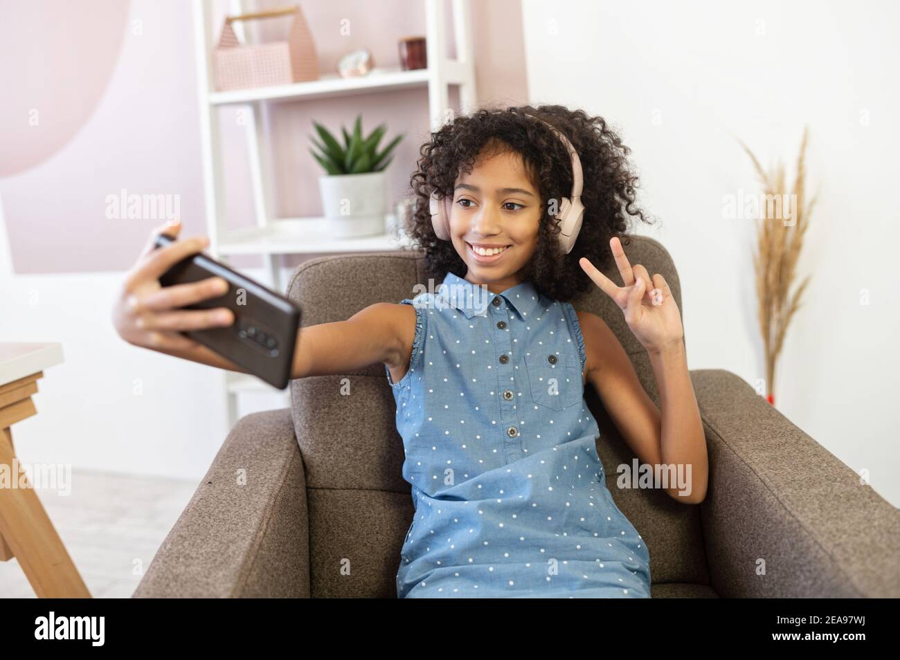 Ein fröhliches, lockiges afroamerikanisches Mädchen mit einem Kopfhörer nimmt ein Selfie auf dem Smartphone auf. Ein hübsches biracial Schulalter Mädchen macht Stockfoto