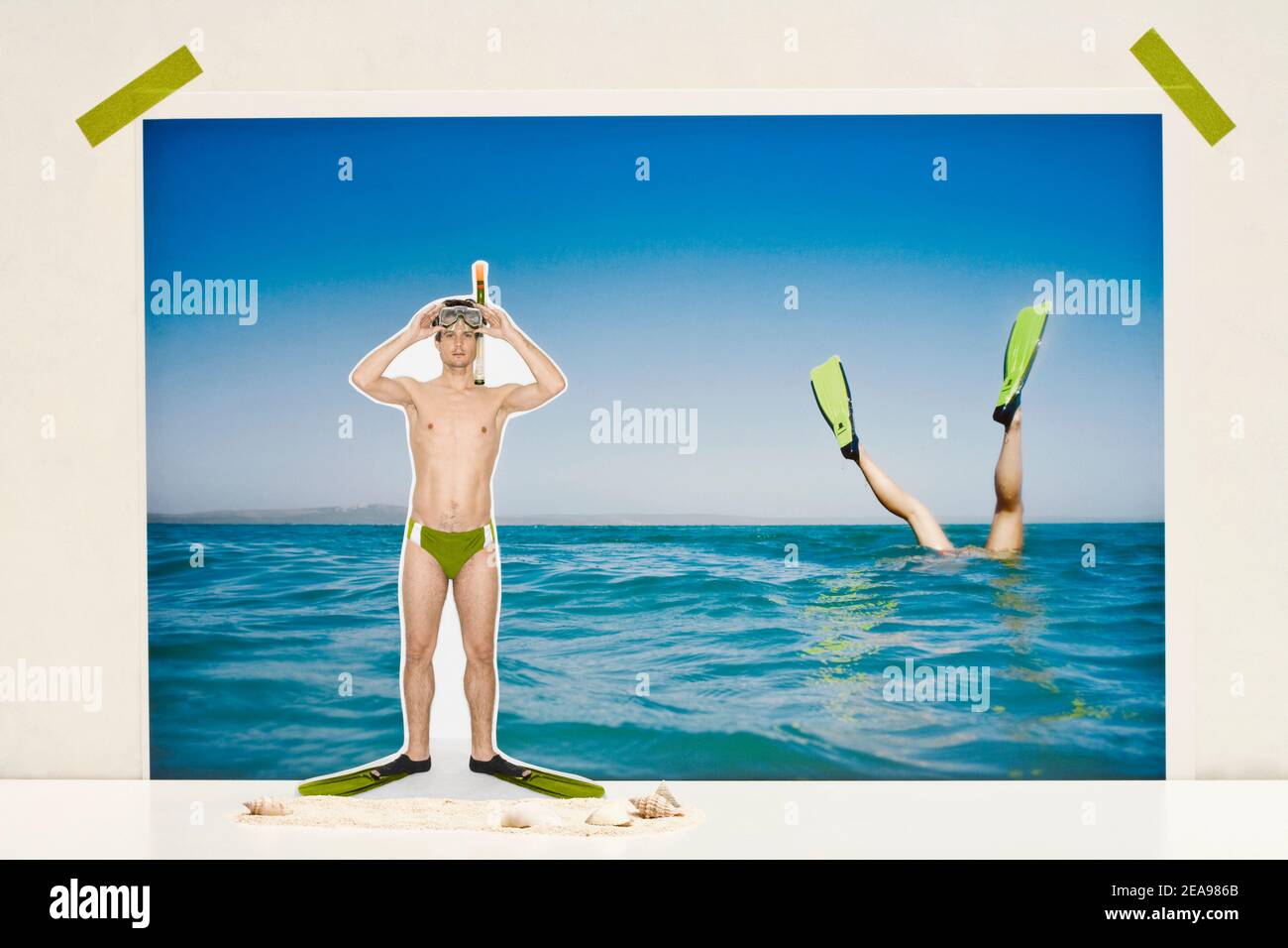 Mann in grünen Badehosen, mit Flossen, Taucherbrille, vor einem Foto des Meeres, mit Sprungbeinen, mit Klebeband fixiert Stockfoto