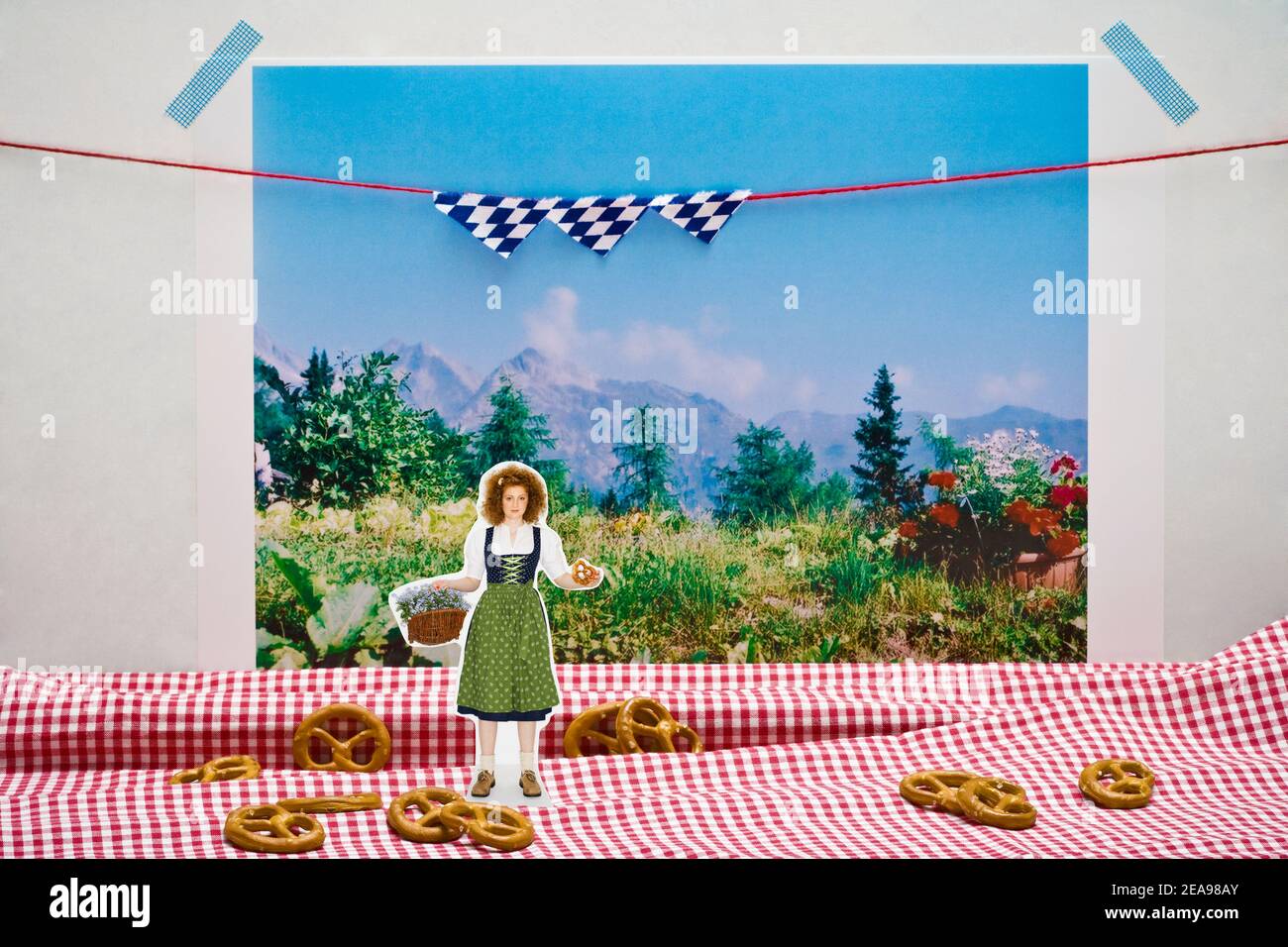 Stillleben Foto, Frau im Dirndl, mit Brezel in der Hand, Blumenkorb, rot-weiß karierte Tischdecke mit Brezeln als Hintergrund, Foto der bayerischen Landschaft als Hintergrund, bayerische Fahnen Stockfoto