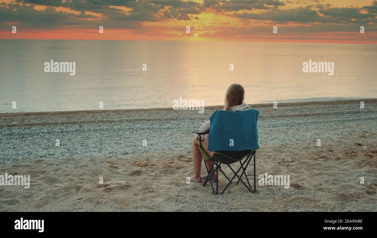 Frau sitzt auf Campingstuhl und trinkt Kaffee. Entspannen und die Natur genießen. Bewundern Sie den Sonnenaufgang auf dem Meer. Stockfoto