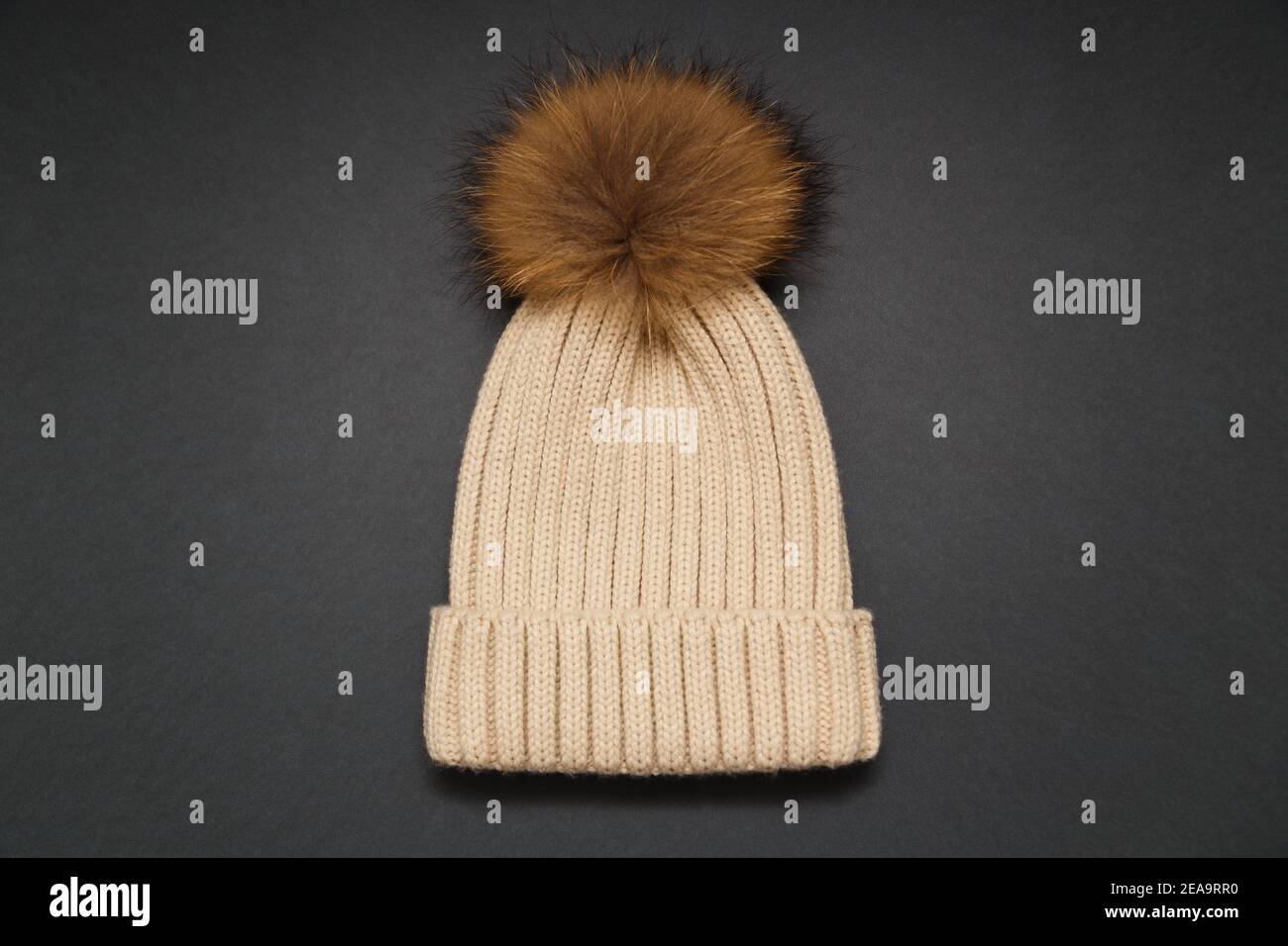 Gestrickte beige Mütze mit Pelzbommel auf grauem Hintergrund. Kuscheliges Strickaccessoire in trendigen Farben, warmes Konzept. Selektiver Fokus Stockfoto