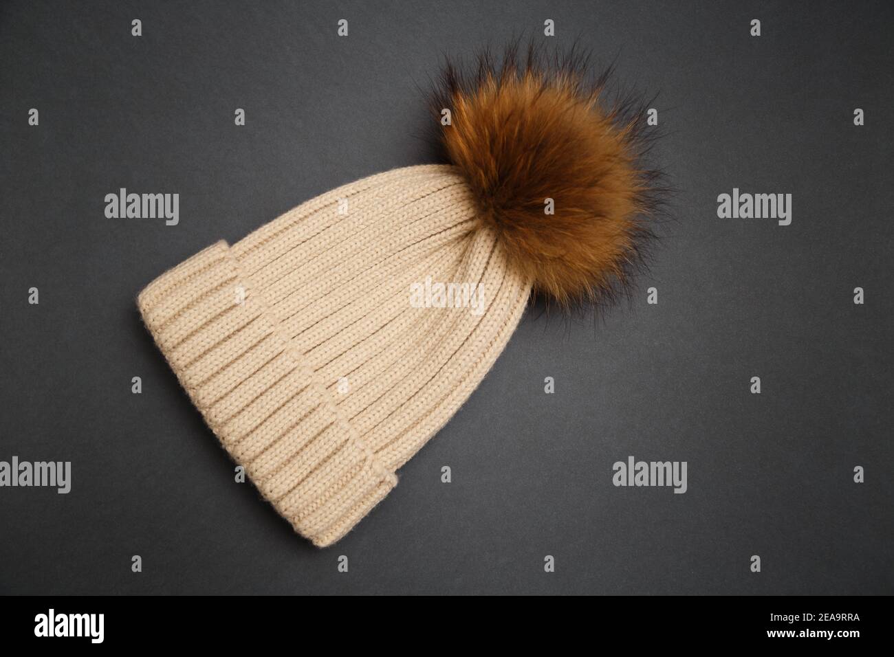 Gestrickte beige Mütze mit Pelzbommel auf grauem Hintergrund. Kuscheliges Strickaccessoire in trendigen Farben, warmes Konzept Stockfoto
