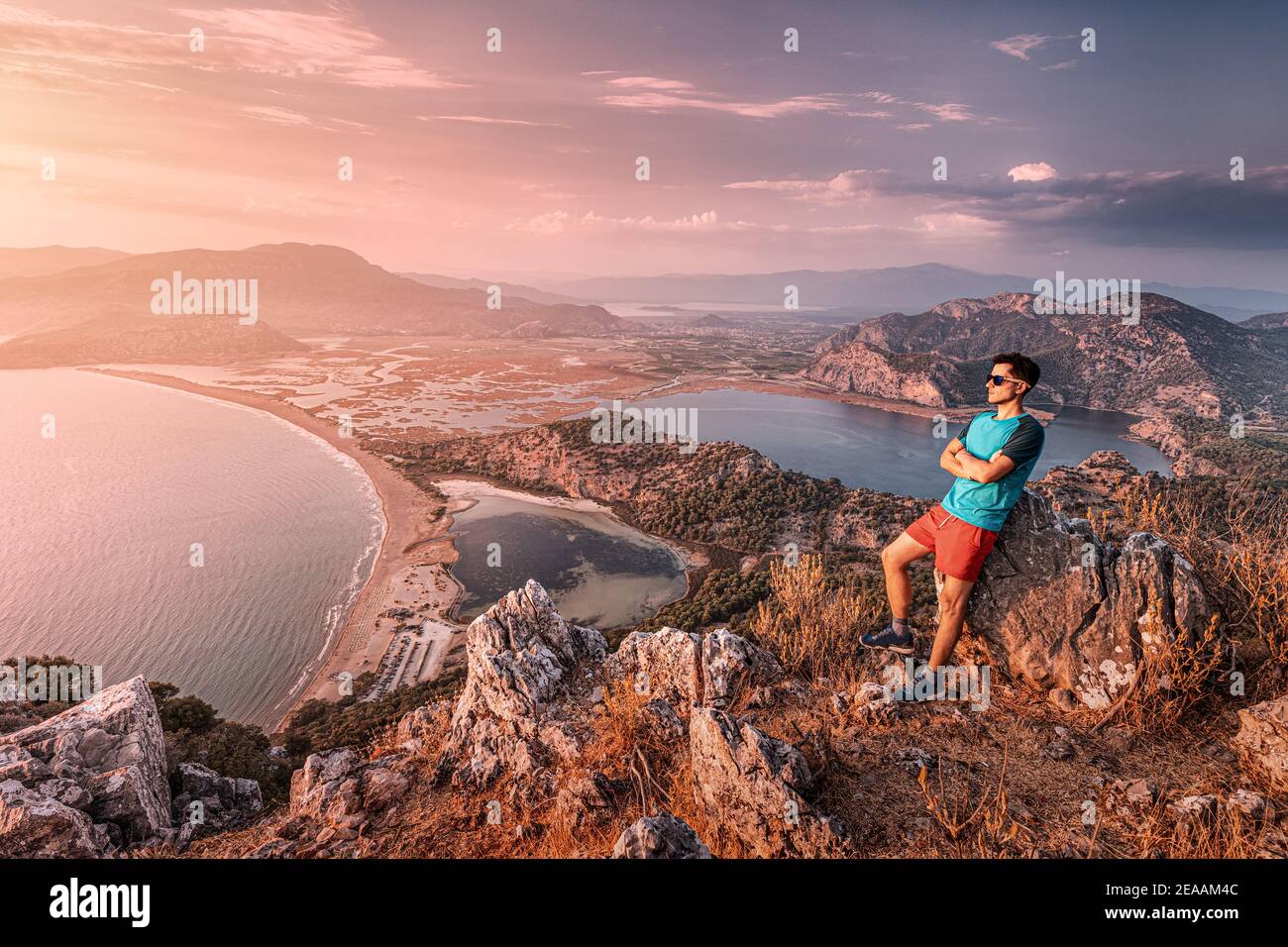 Ein selbstbewusster Mann in Sonnenbrillen und roten Shorts überblickt die Umgebung von einem Aussichtspunkt aus. Im Hintergrund ein farbenfroher Sonnenuntergang über Iztuzu Strand und Stockfoto