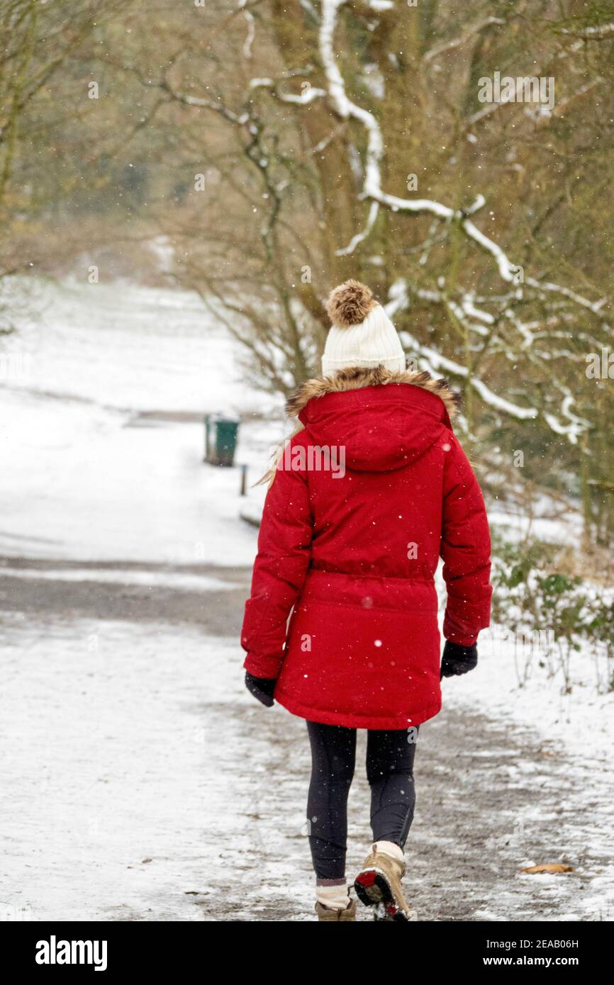 Eine Frau, die einen roten Wintermantel und einen weißen Bommelhut trägt, geht durch den Wald im Schnee. Hampstead Heath, London, Großbritannien. Stockfoto