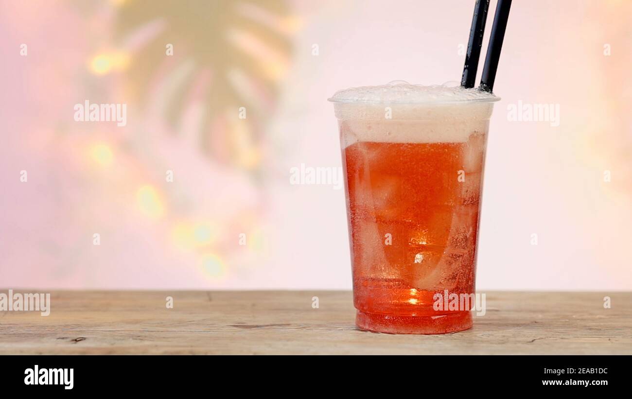 Frisches, pinkes Erfrischungsgetränk in Kunststoff-Take-Away-Tasse mit Eiswürfeln, entkocktem Palmblatt und goldenen Lichtern im Hintergrund Stockfoto