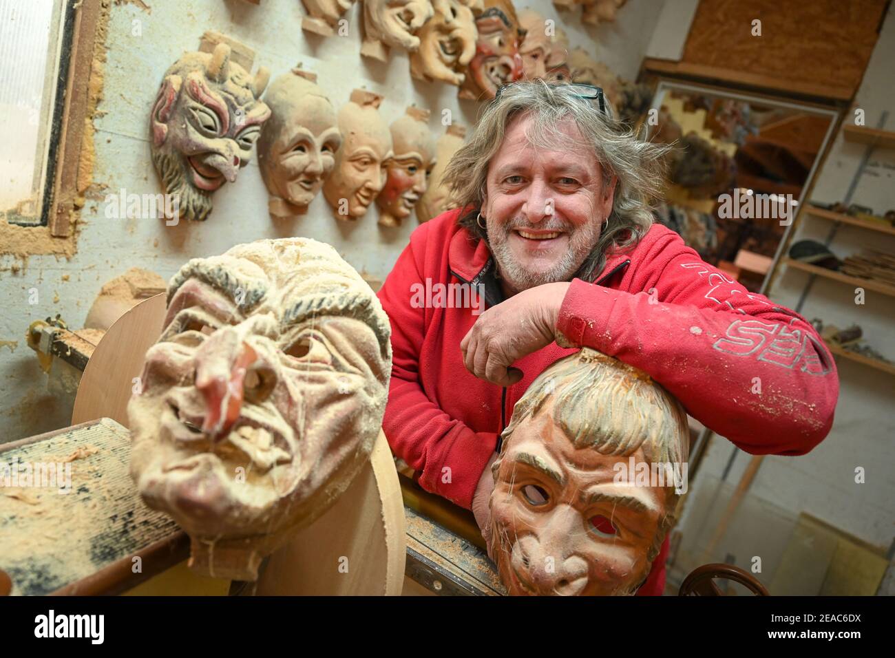 Ravensburg, Deutschland. Februar 2021, 06th. Jogi Weiß, Maske und  Holzschnitzer, lehnt sich in seiner Werkstatt an eine Maske, an der Wand  neben ihm hängen Masken, bedeckt mit Holzstaub und Spinnweben.  Corona-konditioniert, dieses