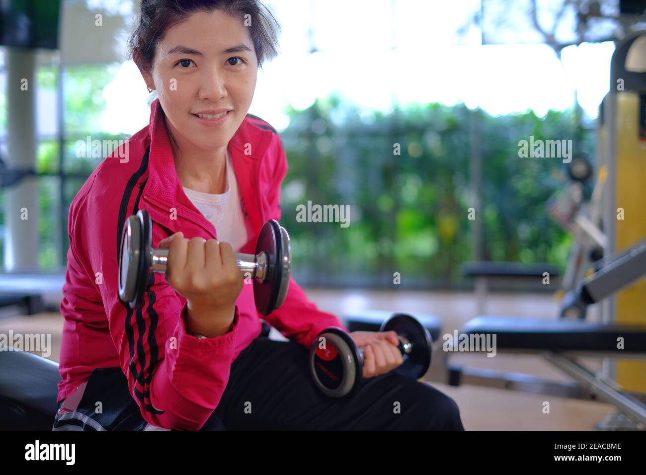 Eine attraktive junge asiatische Frau, die in einem Fitnessstudio arbeitet, Gewicht mit Hanteln hebt, sich müde, aber glücklich fühlt und lächelt. Stockfoto