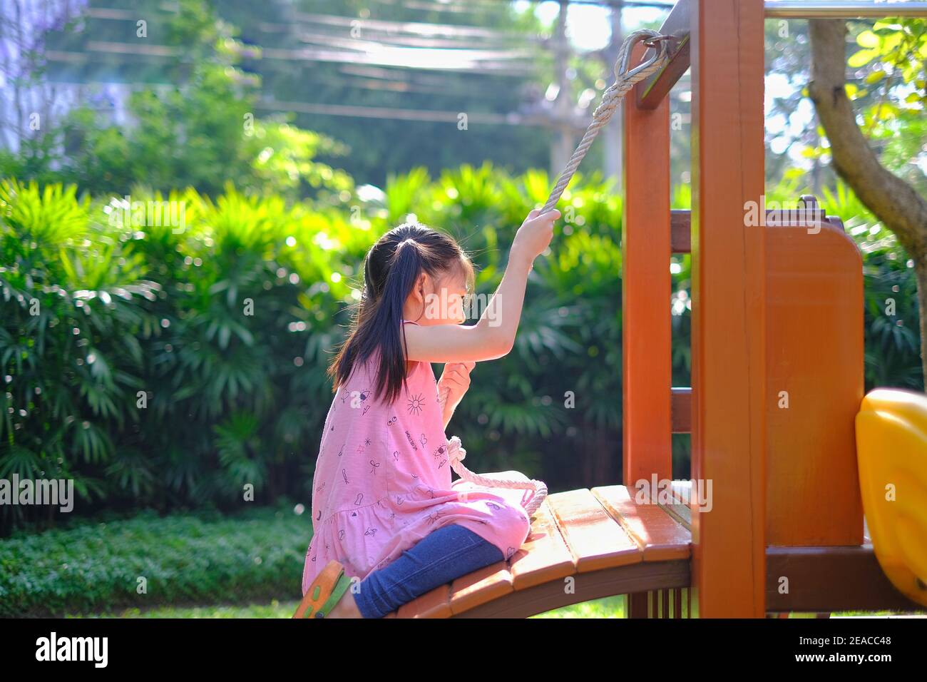 Ein süßes junges asiatisches Mädchen, das eine Holzleiter mit Kletterseil auf einem öffentlichen Spielplatz mit grünem Gras und Baum und einem sonnigen Tag spielt. Stockfoto