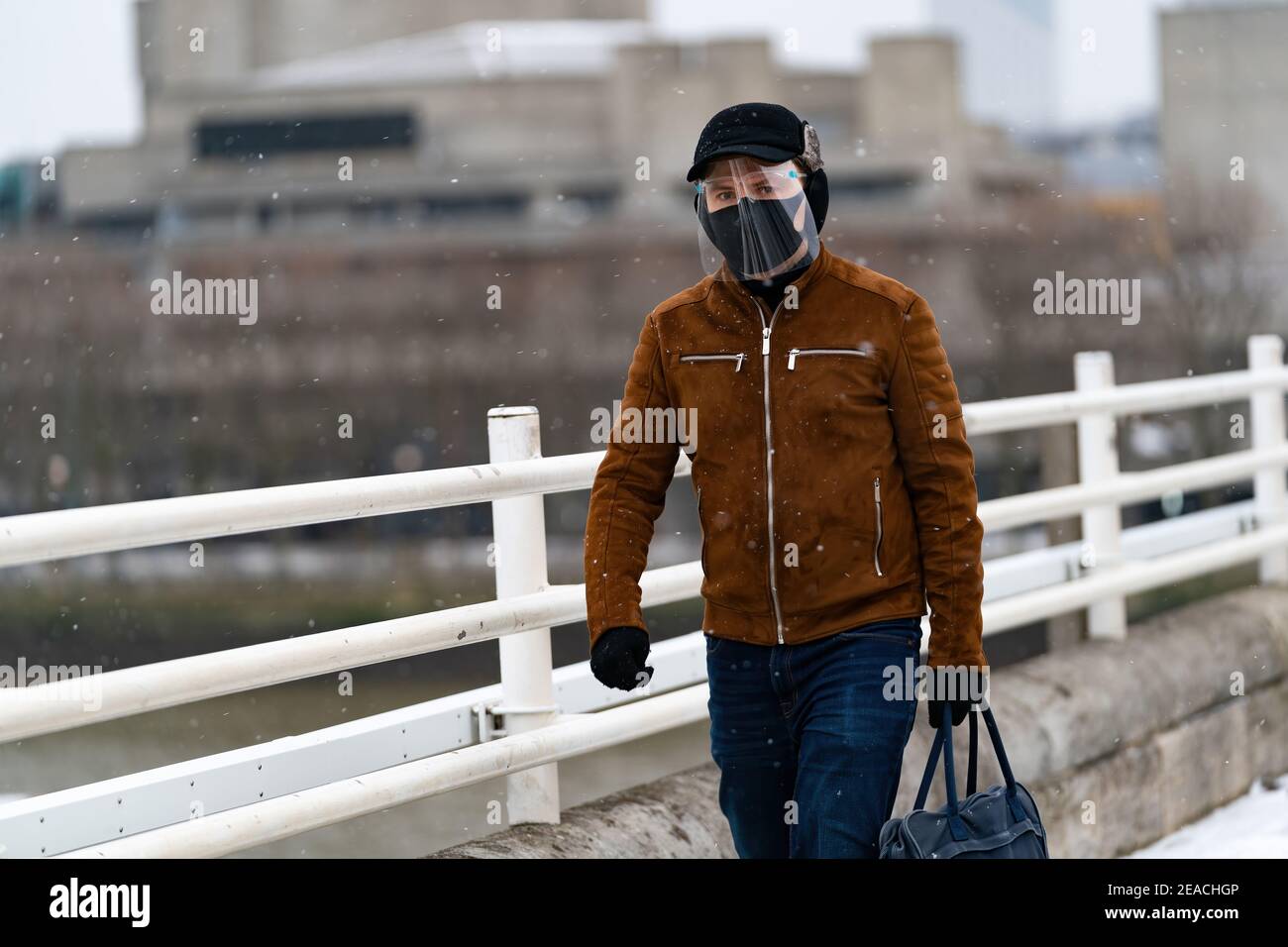 LONDON, ENGLAND - FEBRUAR, 2021: Junger Mann, der die Kamera mit Gesichtsmaske und Gesichtsschild anschaut, läuft auf der Waterloo Bridge in London während eines s Stockfoto