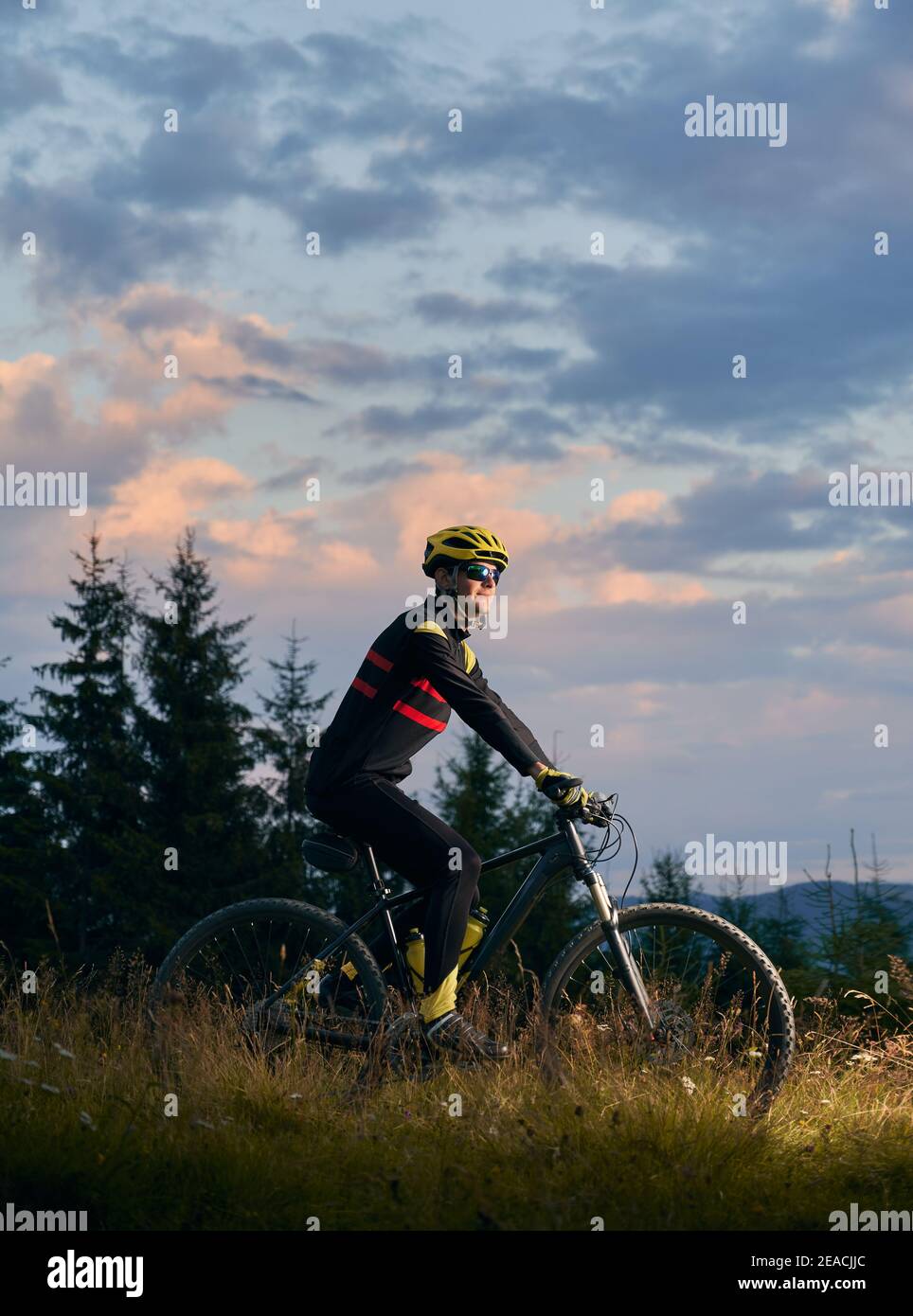 Junger Mann Radfahrer Fahrrad auf Sonnenuntergang in den Bergen. Fahrradfahrer trägt Helm, Sportbrille und Uniform. Seitenansicht. Abendhimmel im Hintergrund. Konzept des aktiven Lebensstils Stockfoto