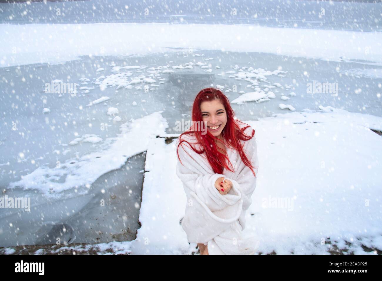Glückliche junge Frau mit langen roten Haaren, nur in einer Decke mit einem fröhlichen Lächeln gewickelt genießt natürliche Kälte-Therapie und Schneedusche im Winter im Freien Stockfoto