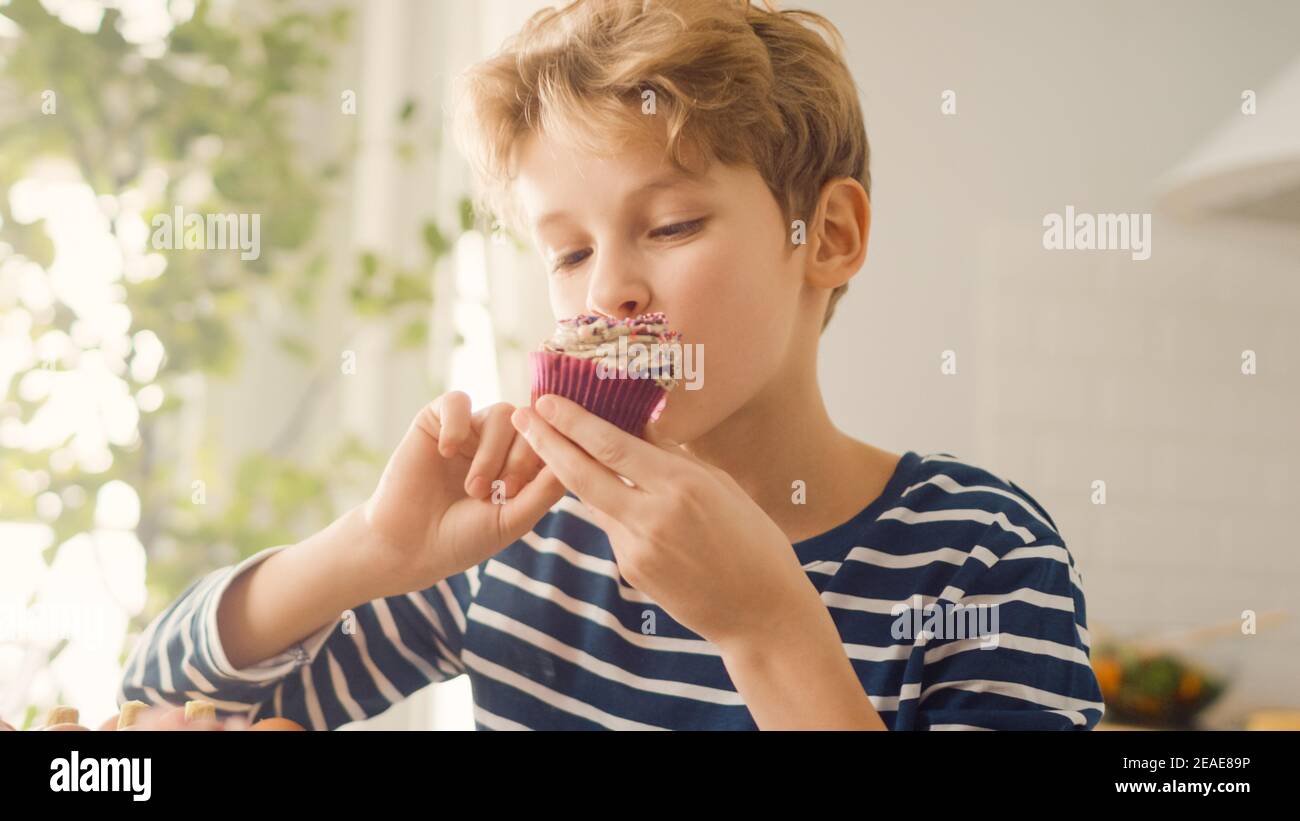 In der Küche: Adorable Boy isst cremigen Cupcake mit Frosting und bestreut Funfetti. Cute Hungry Sweet Tooth Child Bites in Muffin mit zuckerhaltigen Stockfoto