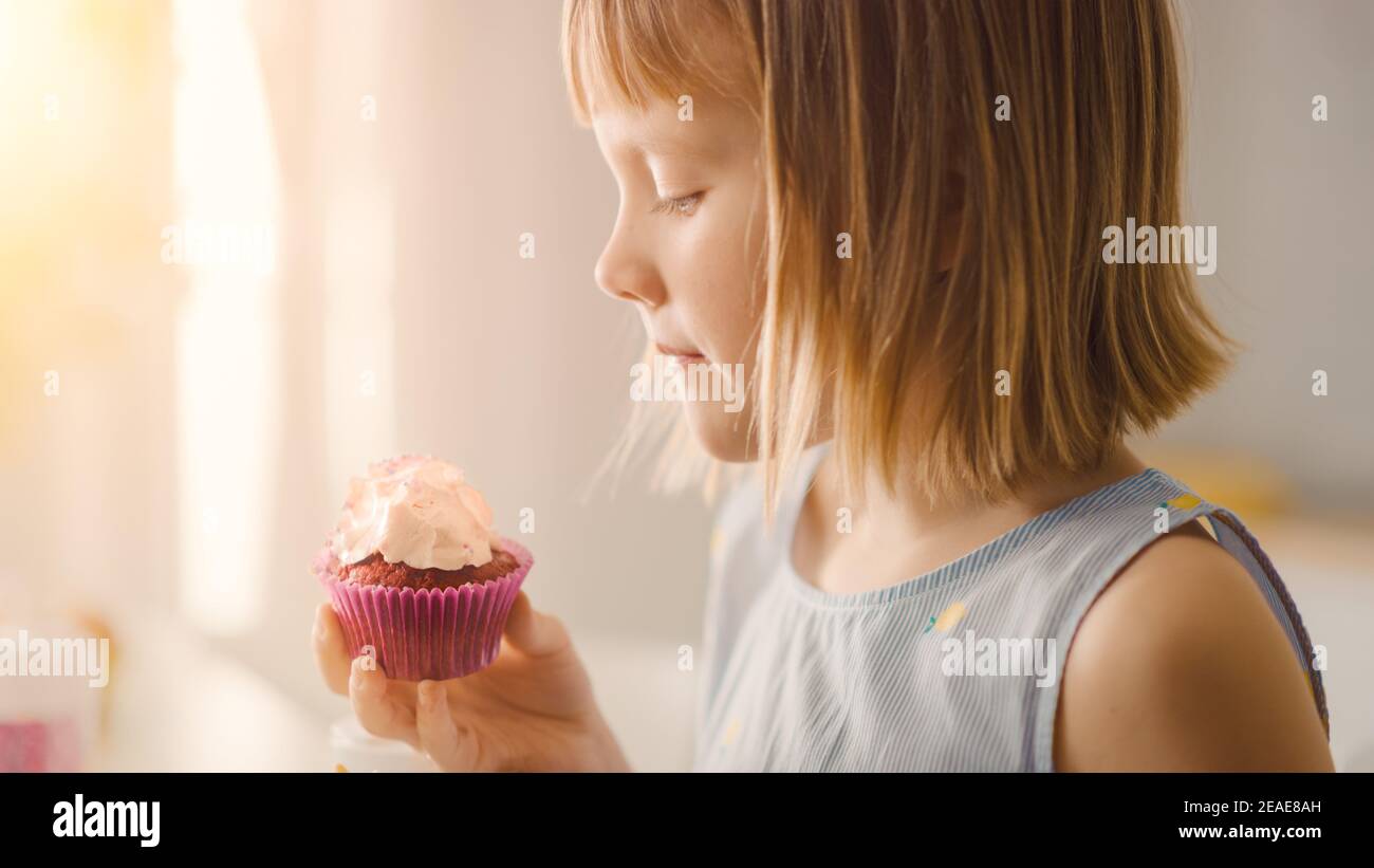 In der Küche: Liebenswert kleines Mädchen isst cremigen Cupcake mit Frosting und bestreut Konfetti. Cute Hungry Sweet Zahn Kind Bites in Muffin mit Stockfoto