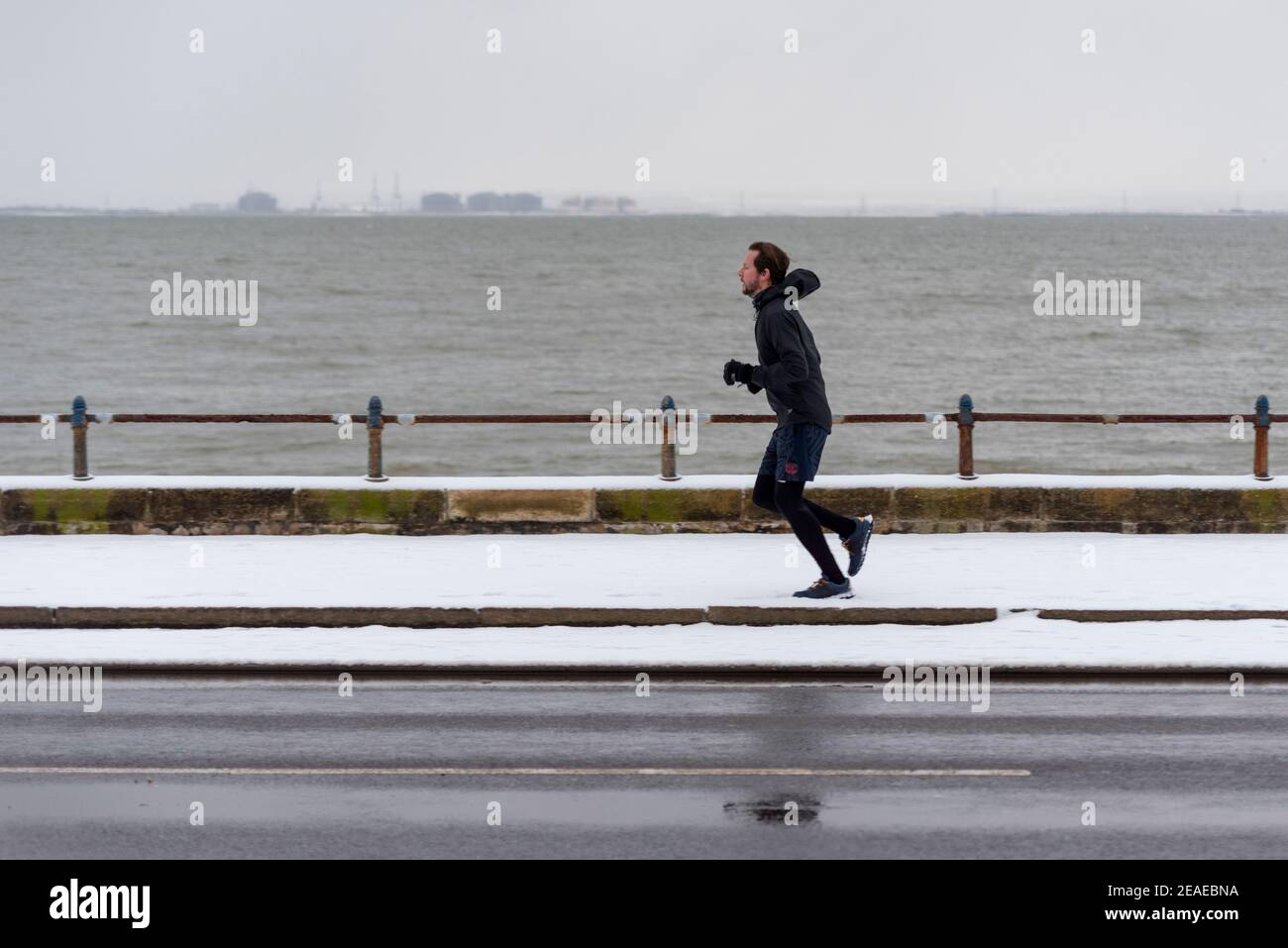 Southend on Sea, Essex, Großbritannien. Februar 2021, 9th. Storm Darcy hat an der Strandpromenade weiteren Schnee fallen gelassen und eisige und windige Bedingungen gebracht. Jogger und Spaziergänger trotzten der Kälte und dem Wind. Rüde Jogger läuft auf Schnee Stockfoto