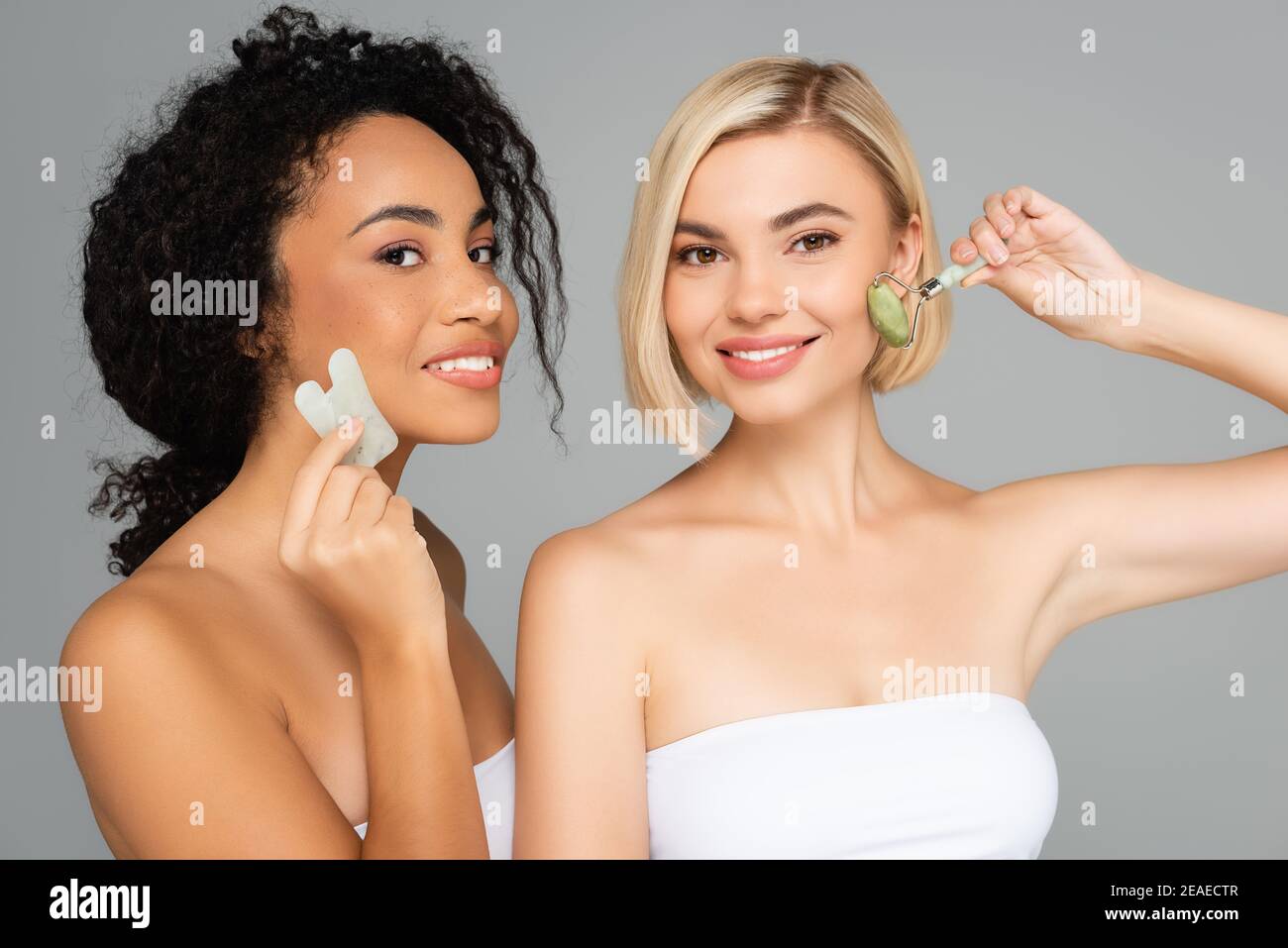 Multiethnische Frauen mit perfekter Haut massieren Gesichter mit Gua Sha Und Jadewalze isoliert auf grau Stockfoto