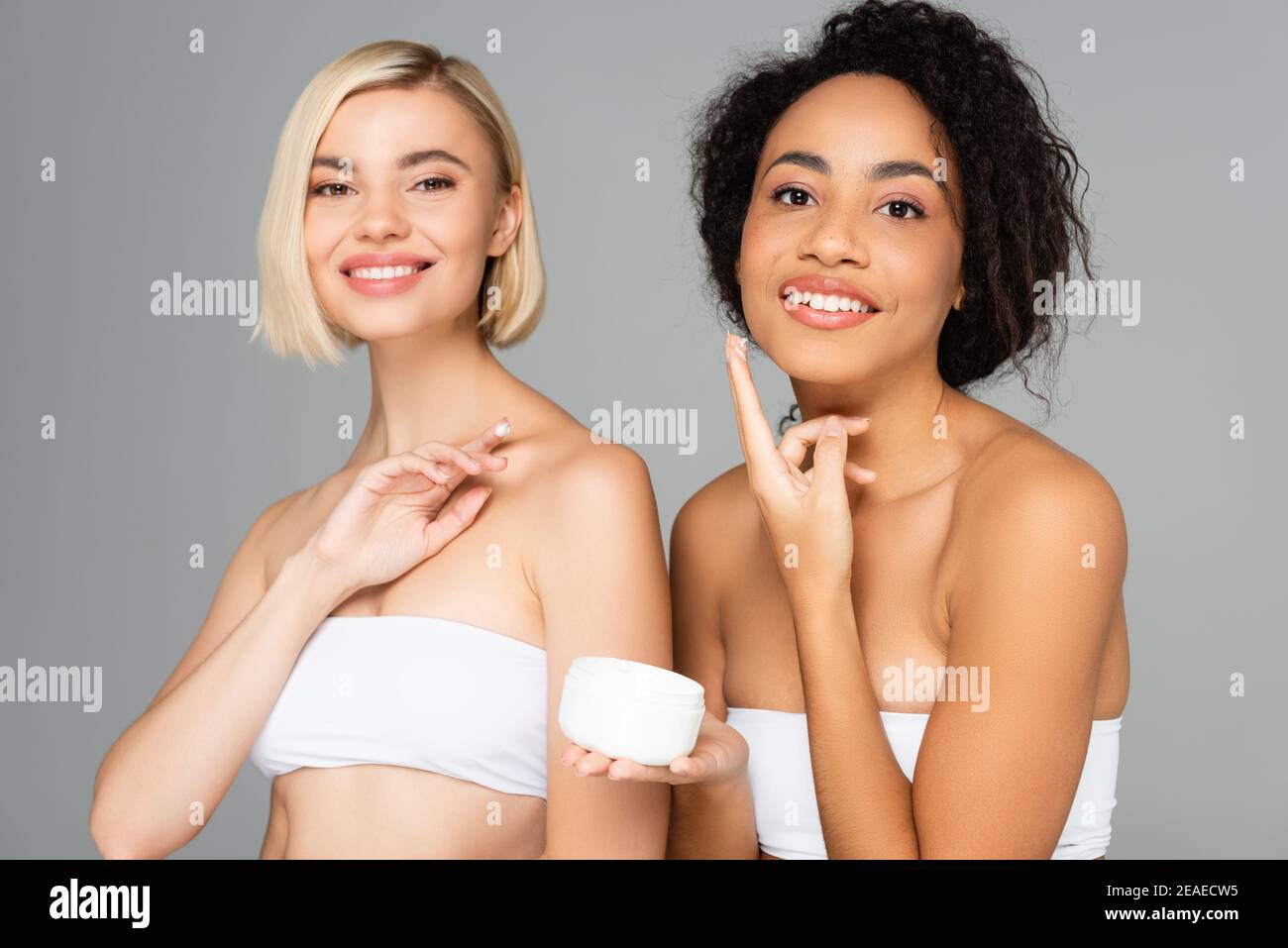 Lächelnd interracial Frauen mit perfekter Haut hält kosmetische Creme isoliert Auf Grau Stockfoto