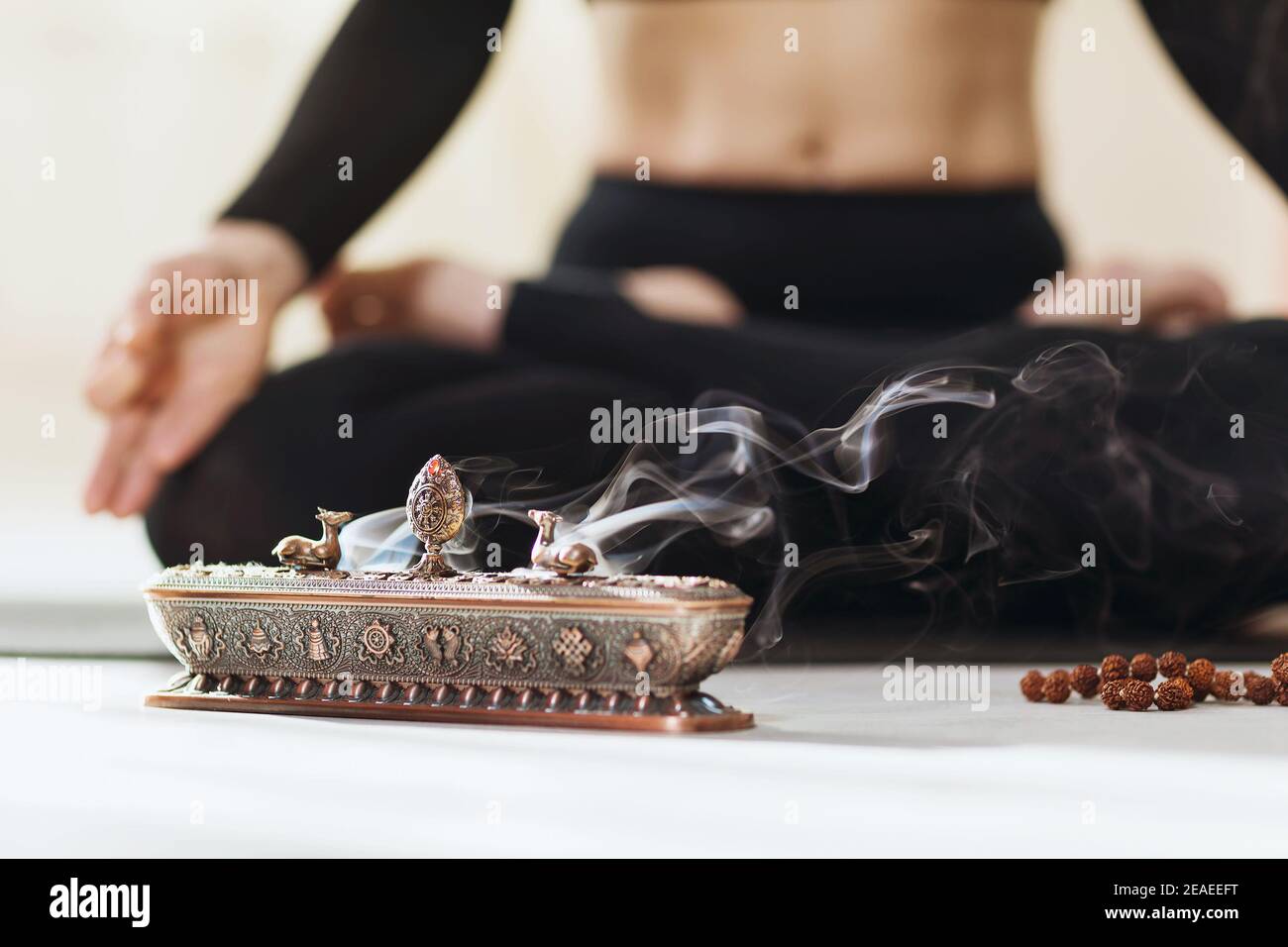 Metallständer für Räucherstäbchen mit Rauch auf dem Hintergrund einer meditierenden Person in der Lotusposition. Stockfoto