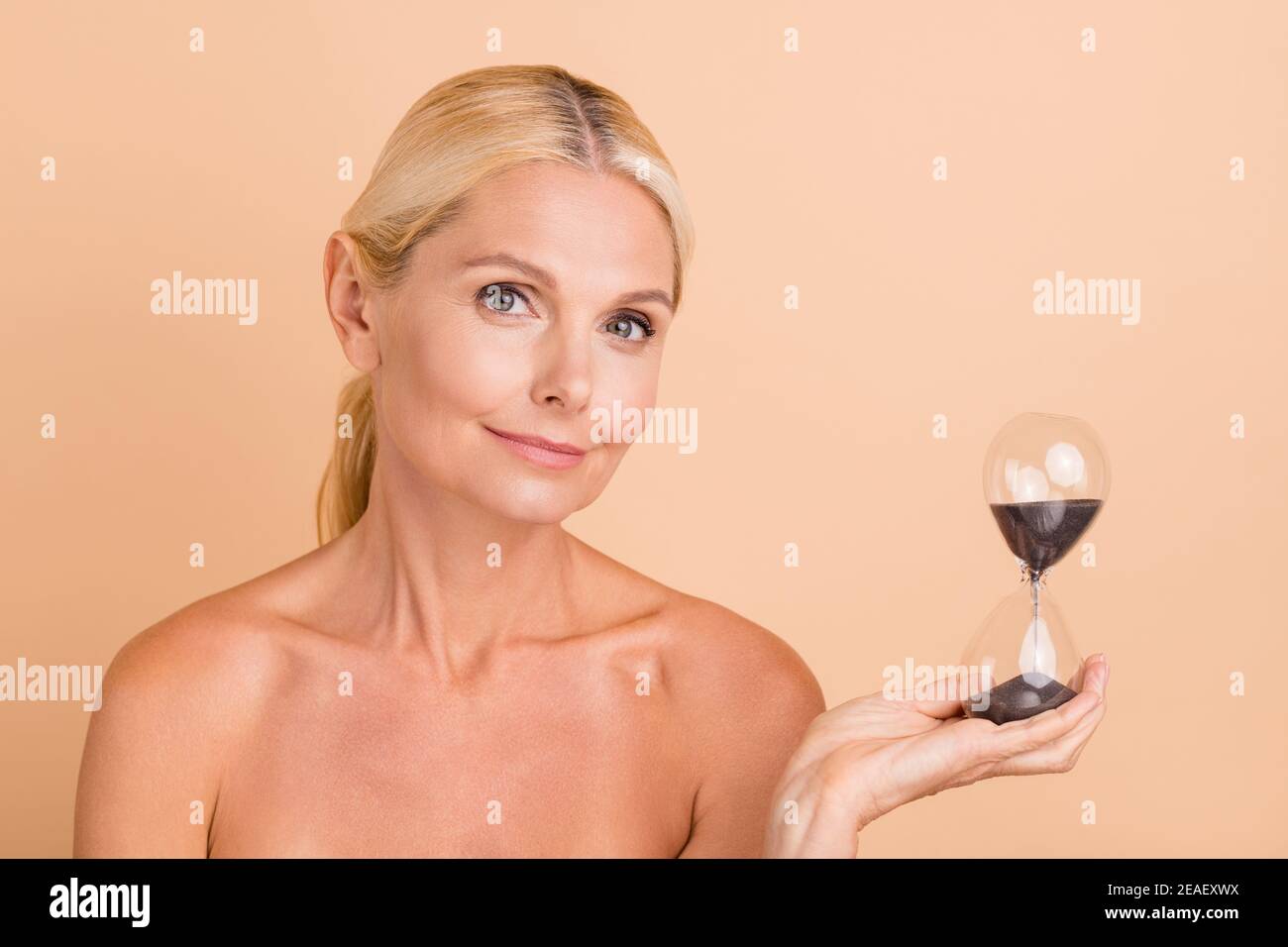 Portrait von charmanten Frau mittleren Alters halten Sand Stunde Uhr Show Time Hautpflege Therapie Verfahren isoliert beige Farbe Hintergrund Stockfoto